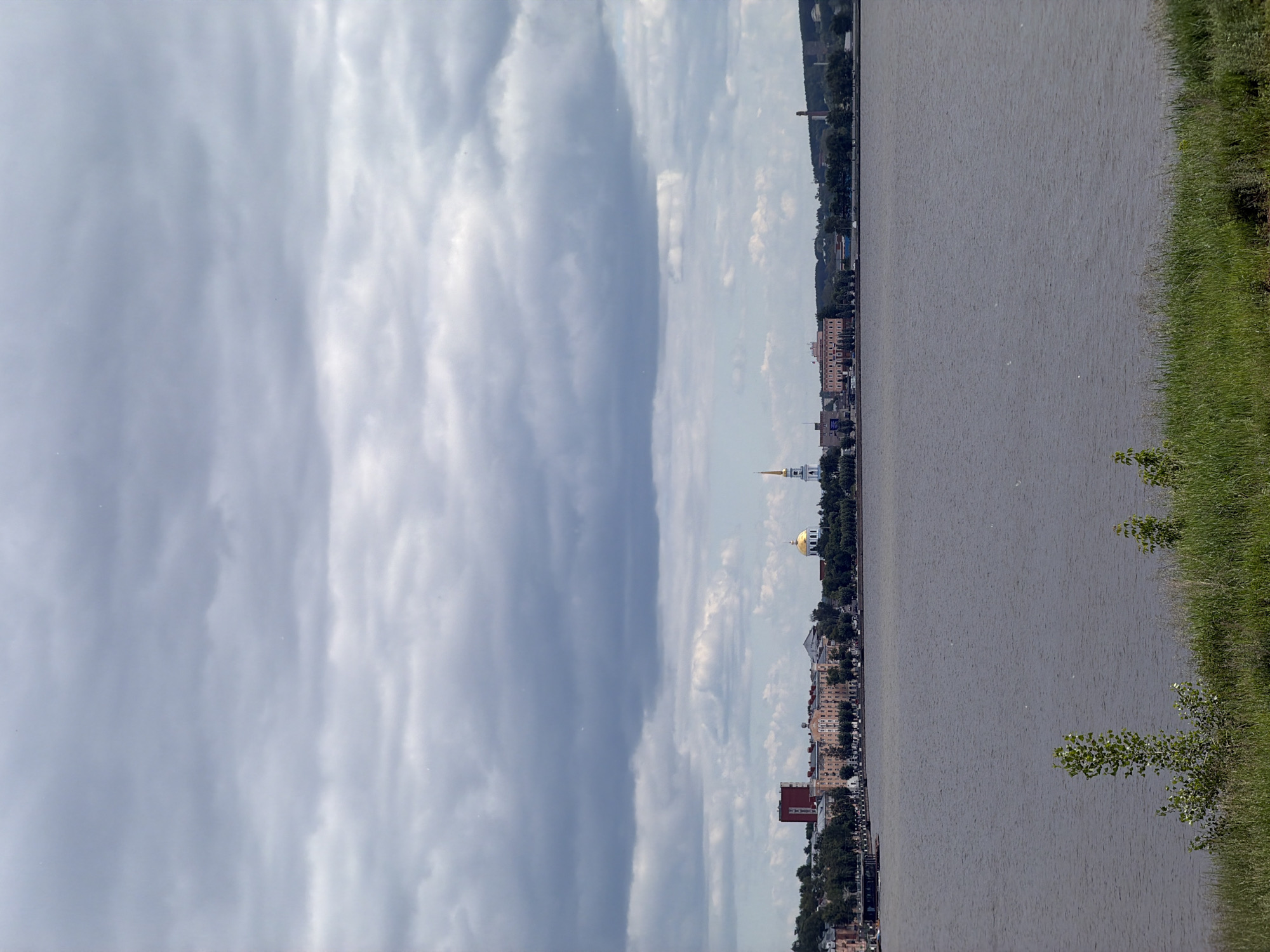 A wide, calm river dominates the foreground and middle ground of the image. The river's surface is a dull gray, reflecting the overcast sky. Lush green grass and small trees line the near bank of the river. In the distance, across the water, lies a city skyline featuring several buildings of varying heights and architectural styles. Prominent structures include a tall, red-brick building, a long, pale pink building with multiple windows, and a white church with a golden dome and a tall spire topped with a cross. The city is bordered by a line of trees, and further in the distance, a smokestack can be seen, indicating industrial activity. The sky above is heavily clouded with large, dark gray clouds, suggesting an overcast or impending rainy day. The overall impression is a serene, yet somewhat somber, urban landscape seen across a broad body of water.