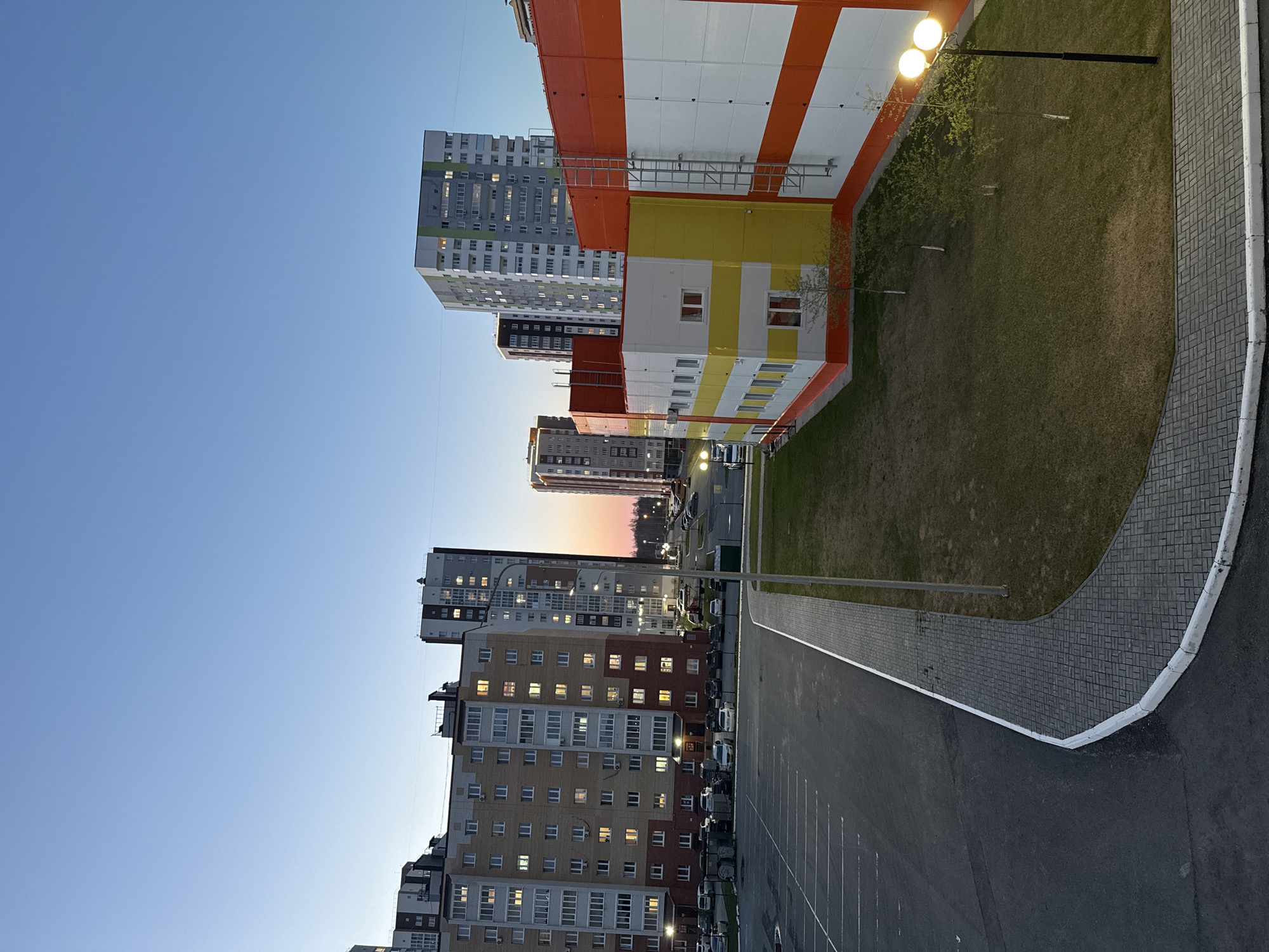 The image shows a residential area in Tobolsk, Russia at dusk. Several apartment buildings of varying heights are visible, some with lit windows. A parking lot is in the foreground, adjacent to a sidewalk and a grassy area with lampposts. The sky is blue, transitioning to orange near the horizon.