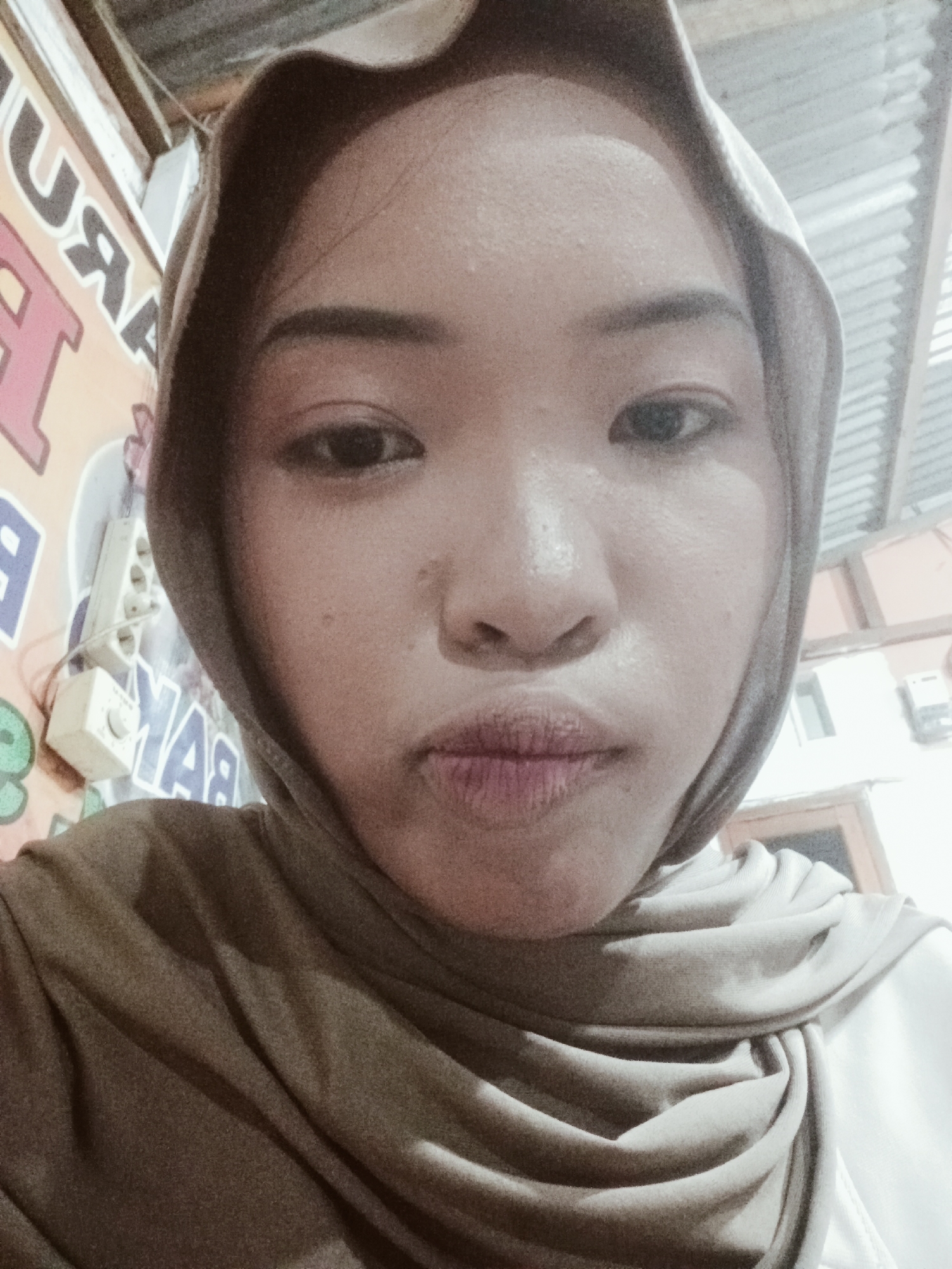 A selfie is taken from a slightly low angle, featuring a young woman wearing a tan hijab. Her face is mostly centered in the frame, with her lips slightly pursed. The background shows a corrugated metal roof, and to the left, there is signage with text that includes letters like "U", "R", and "A". Partially visible on the wall to the left is a white electrical outlet with three sockets and a dial. The overall lighting suggests an indoor setting, likely during the daytime, with shadows cast by the roof structure. The image quality is moderate, with visible grain. The specific location is within the city of Bumbung, Indonesia.