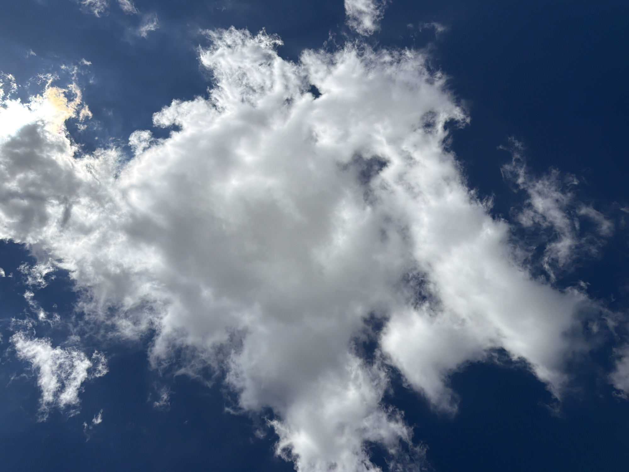 A low-angle, vertical, full-frame shot captures a deep blue sky filled with a variety of clouds. A large, dense cumulus cloud dominates the center of the frame, its white mass displaying varying shades of gray, indicating significant depth. To the upper right, a cluster of smaller, wispy clouds is illuminated by the sun, which is partially visible through the haze, casting a yellowish-orange glow. The rest of the sky is a rich, dark blue, with more scattered, thin clouds appearing as white streaks in the upper left and lower sections of the image. The scene is devoid of any human presence, structures, or discernible geographical features, offering a pure vista of the sky.