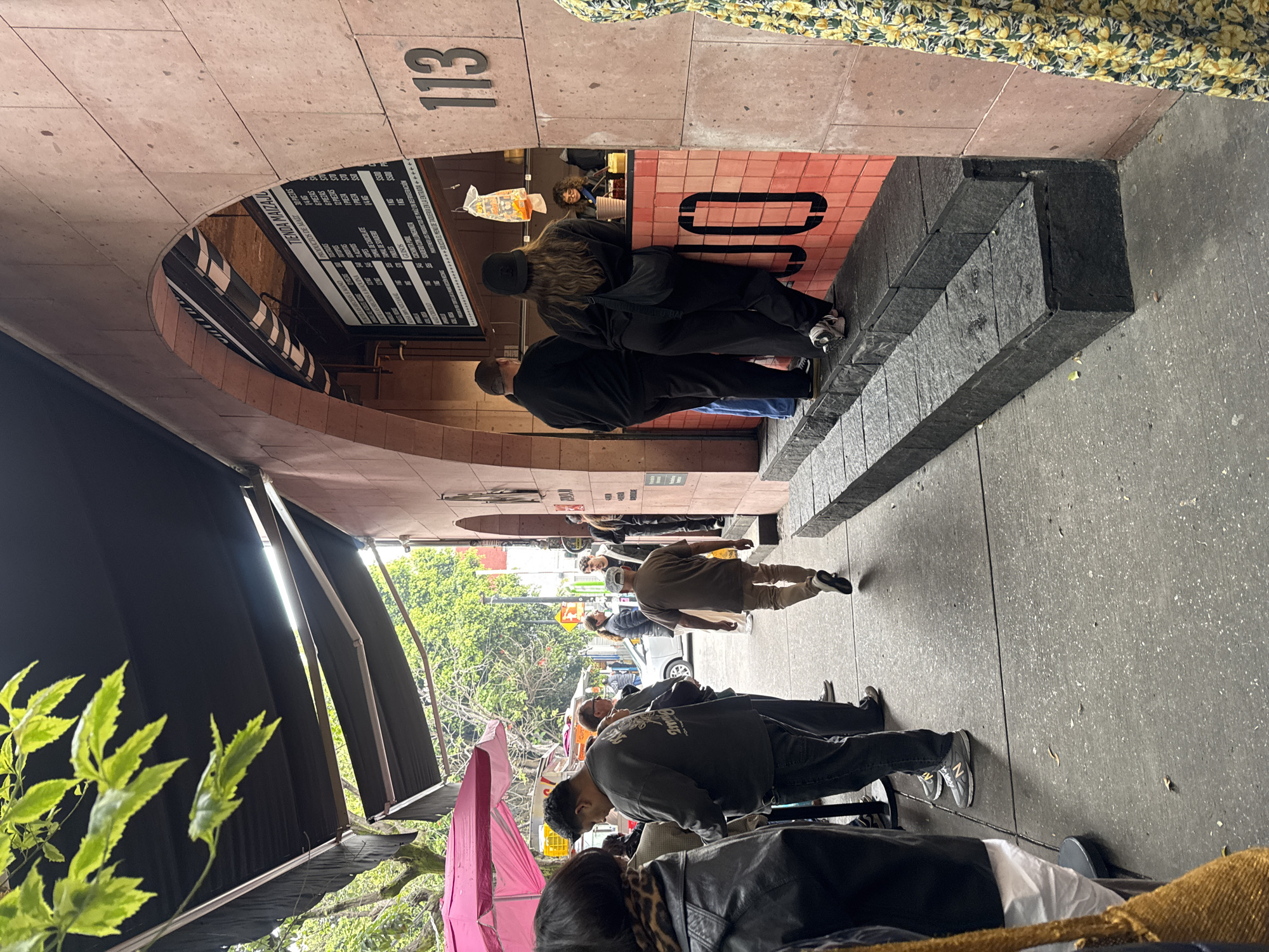 Here is a detailed description of the image:
**Scene:**
The image captures an urban street scene in Mexico City, Mexico. The focus is on the entrance of a taqueria named "Tienda Maizal," located at address "113". People are queuing to order food from the small restaurant.
**Environment:**
The taqueria is built into a building with red-tiled archways. The restaurant front has a menu board displayed. On the sidewalk, there are street vendors with a pink umbrella, and people are walking by. Green trees and other buildings are visible in the background.
**People:**
Several people are lining up at the taqueria, either placing orders or waiting for their food. Other people are passing by on the sidewalk.
**Objects:**
* Menu board with the price of Mexican food.
* Pink umbrella.
* Plant leaves.
* Street furniture.
**Actions:**
People are queuing, walking, ordering food, and going about their daily activities.