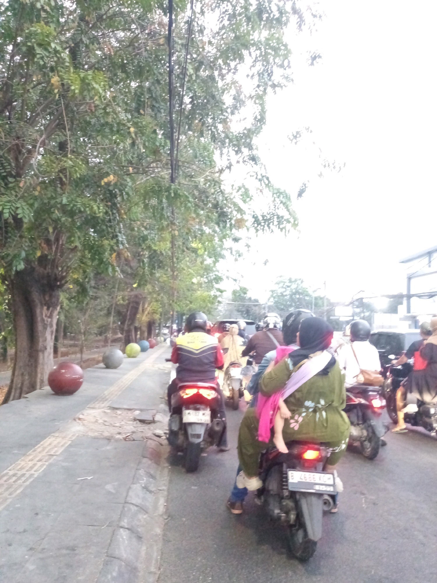 The image captures a street scene in Bekasi, Indonesia, likely during the day given the bright sky. A line of trees borders a sidewalk, on which several large, spherical decorative objects are placed, including one red, one grey, and one yellow sphere. The sidewalk is paved with textured tiles and concrete. A man wearing a yellow and black vest is riding a motorcycle on the road, with the back of his head and the motorcycle's taillights visible. Behind him, several other motorcycles and riders are visible, some with passengers. One motorcycle in the foreground has a child sitting on the lap of a woman who is wearing a green garment with floral patterns and a pink wrap. The road is asphalt and appears to be in moderate traffic. The sky is overcast or very bright, suggesting a hazy or cloudy day.