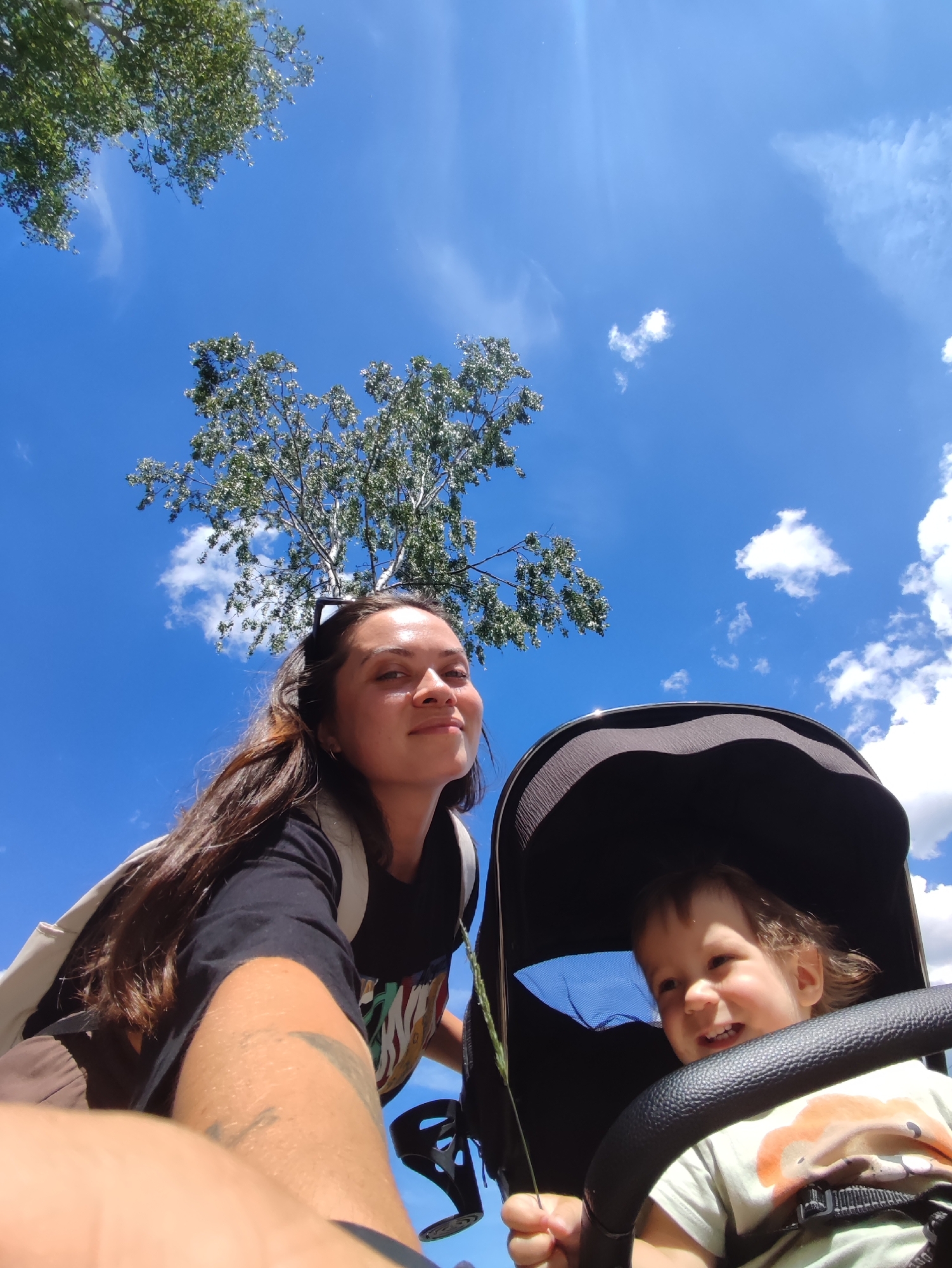 This is a low-angle, selfie-style shot taken outdoors under a bright blue sky with scattered white clouds. A young woman with long, dark hair is in the foreground, leaning forward and looking at the camera. She is wearing a black t-shirt and a light-colored backpack. To her right, a baby is visible in a stroller, looking to the side and smiling. The baby is wearing a light-colored t-shirt with a graphic print. Trees with green foliage are visible in the background, with branches reaching into the sky. The overall scene suggests a casual outdoor activity, possibly a walk in a park. The image does not contain any elements that would specifically indicate it was taken in the city of Chaikovsky, Russia.