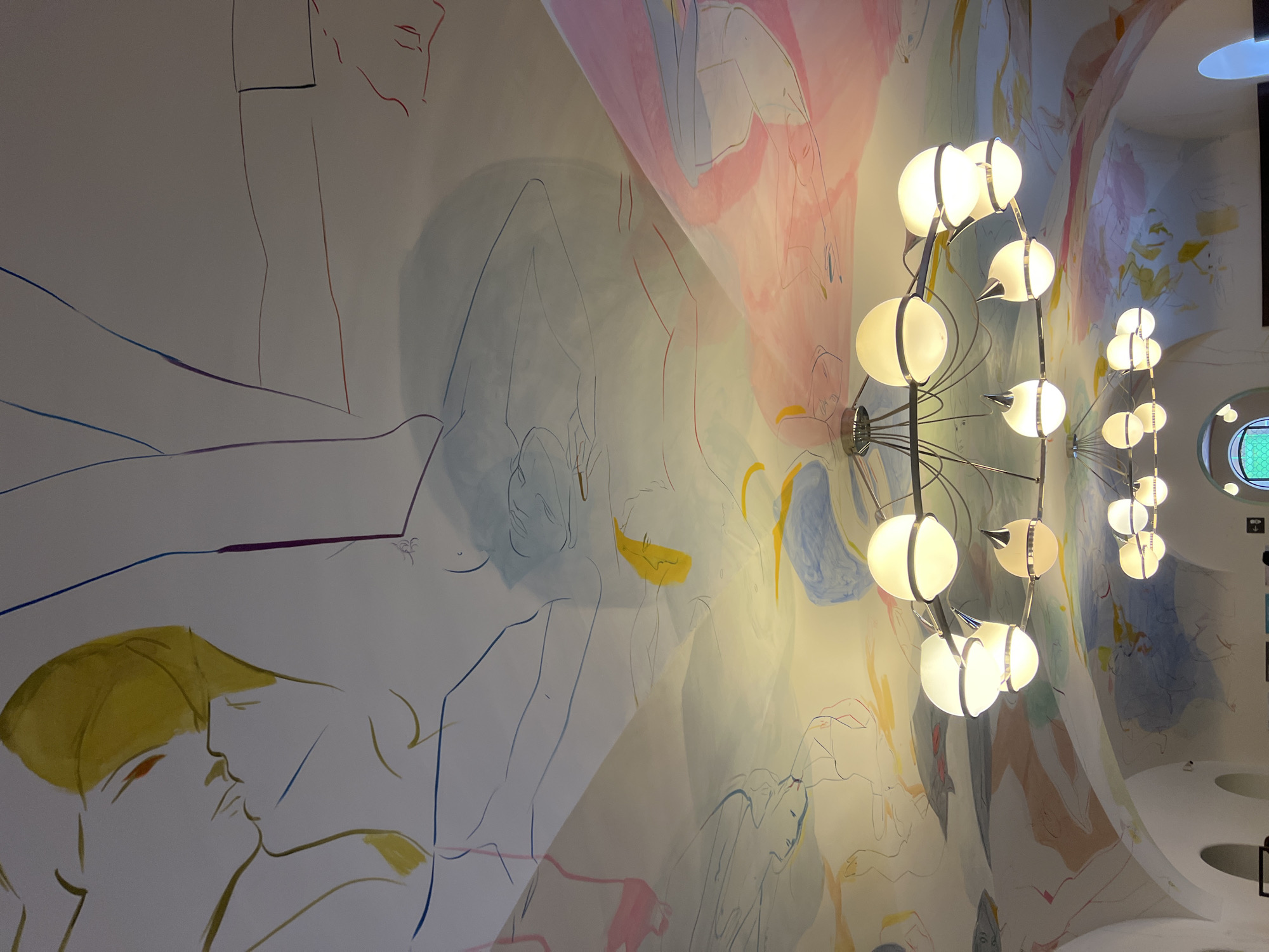 This media file presents an immersive view of a unique interior space, likely in London, United Kingdom, characterized by a large-scale mural painted across a dramatically curved wall and ceiling. The artwork features numerous stylized and fluid line drawings of human figures, some abstract, others more clearly discernible, often depicted in various reclining or intertwined poses. These figures are rendered with minimalist outlines, accented by soft, watercolor-like washes of color in shades of pink, blue, yellow, and grey against a light background. One close-up detail reveals two figures with yellow head coverings, appearing to kiss.
Illuminating this artistic environment are multiple contemporary light fixtures mounted along the curved surface. Each fixture comprises several spherical or semi-spherical white globes, emitting a warm, inviting glow from their sleek metal frames, casting soft light and shadows across the mural.
In the distance, a circular window or opening with a green, grid-like pattern offers a glimpse into an adjacent area or the exterior. The overall aesthetic suggests a sophisticated and art-infused setting, possibly found in a modern hotel, gallery, or a public space designed for aesthetic contemplation and a sense of human connection. The image is a static capture of this distinctive interior, with no active event or specific activity taking place. No discernible text is visible within the frame.