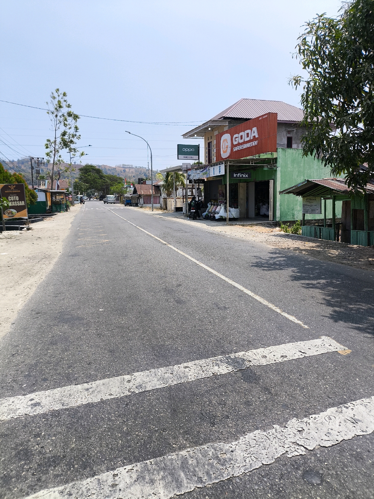 The image shows a street scene in a city in Indonesia. The road is asphalt and has white dashed lines for lane markings. There are buildings on the right side of the road, including shops with signs for "GODA SAFERSMARTEV", "OPPO", and "Infinix". One building is painted green and has a small covered seating area in front of it. On the left side of the road, there is a sign for "COTO WASSAR". There are some trees and utility poles visible, and a few vehicles are on the road in the distance. The sky is clear and blue. The scene appears to be during the daytime.