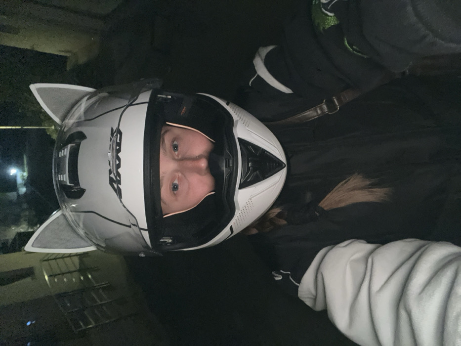 A single individual is depicted in a close-up, self-portrait style photograph, captured at night in Rafailovići, Montenegro. The subject is wearing a white AROX brand motorcycle helmet, which features two grey, semi-translucent, cat-ear shaped attachments on its upper surface. The helmet's clear visor is raised, exposing the subject's face. Visible facial features include light-colored eyes and a small piercing in the right nostril. Brown hair, appearing to be braided, extends from beneath the helmet on the subject's left side. The individual is dressed in a dark jacket, with a lighter-colored sleeve visible on the left arm. A brown strap, consistent with a shoulder bag, crosses over the subject's right shoulder. The background is dark and indistinct, illuminated by faint, blurry light sources, and features subtle outlines of a building with railings on the left, indicative of an outdoor, nighttime urban or residential setting.