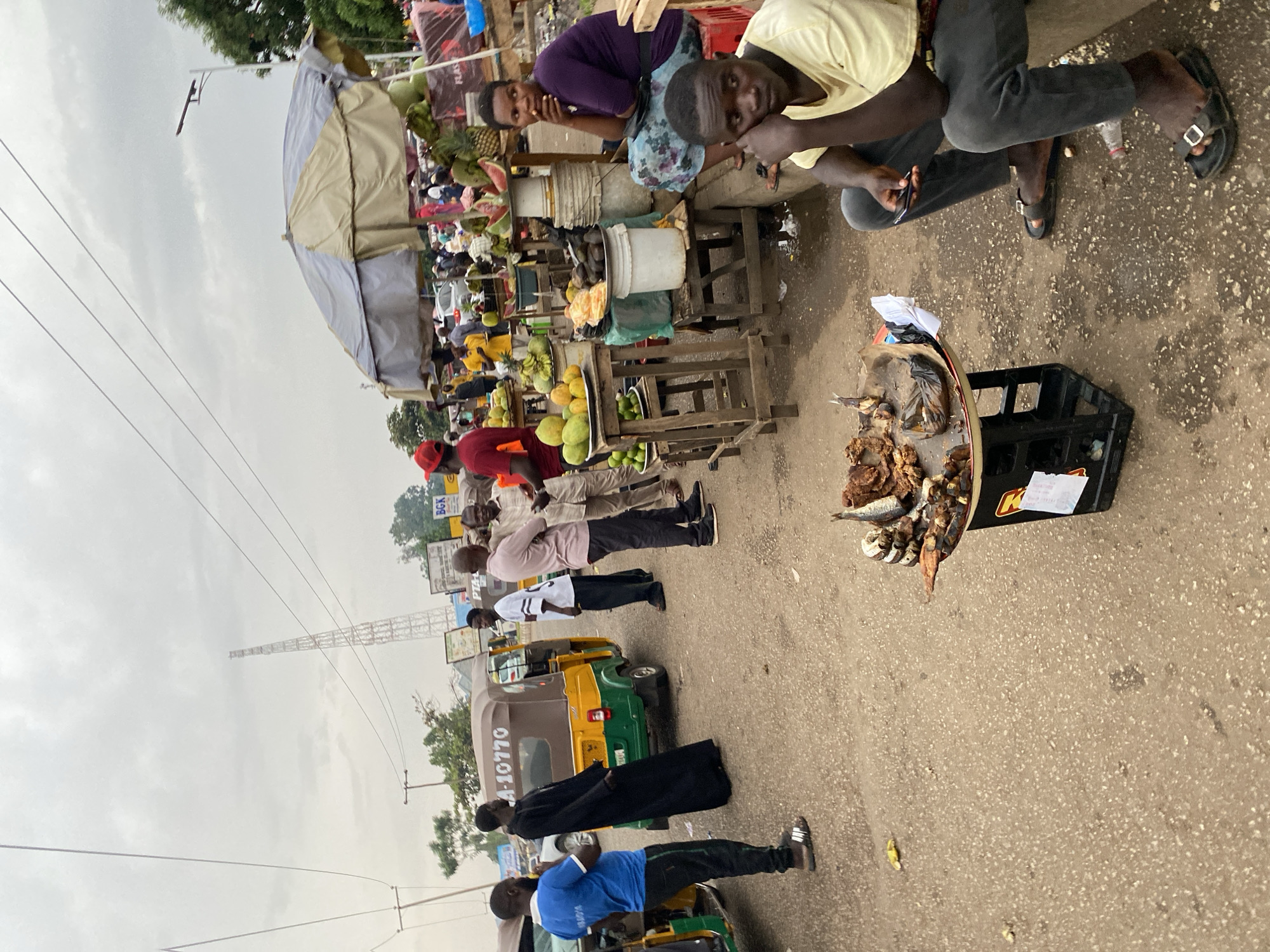A street scene in Bukuru, Nigeria, shows a busy market with several vendors and passersby. Two people are positioned near the top of the frame, likely selling fruits under an awning-covered stall. The adjacent stalls have various fruits displayed, including mangos, pineapples, and watermelons. Further down the road, other people walk along the roadside, next to yellow and green tricycles with the number "A-10770" visible on one. A tower is visible against the cloudy sky in the background. In the foreground, a tray of dried fish sits on top of a black crate on the pavement near a person sitting on a stool.