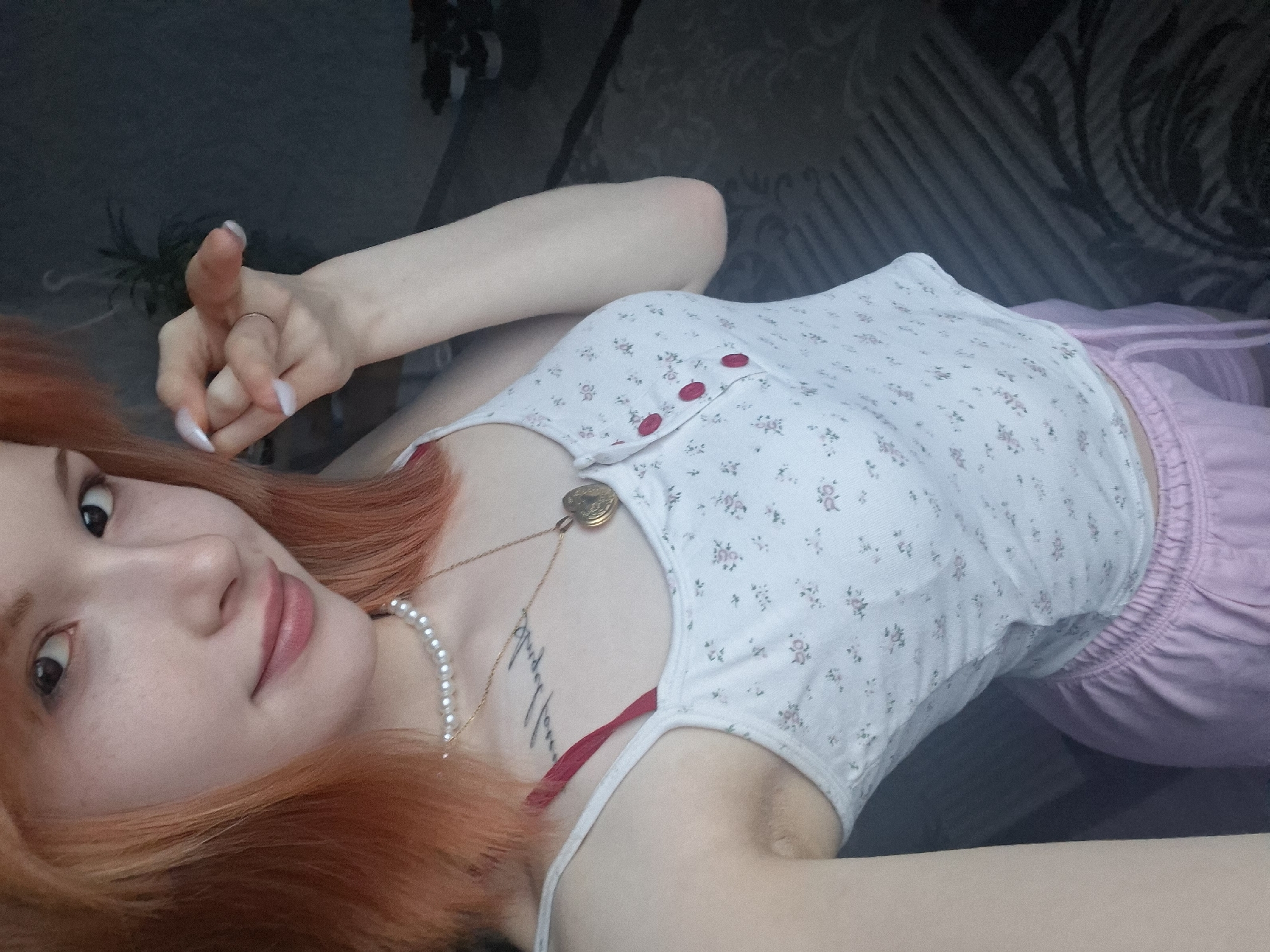 The image is a high-angle, close-up portrait of a young woman lying down indoors, likely in Chaikovsky, Russia. She has fair skin, striking reddish-orange hair, and dark eyes, and she looks directly at the camera with a gentle, relaxed expression and a faint smile.
The woman is wearing a white tank top with a subtle floral pattern of small pink and green flowers, accented by three red buttons down the front. She also wears light pink shorts with an elasticated waistband. Her accessories include a gold chain necklace with a small, heart-shaped locket and a pearl necklace. A vertical, stylized tattoo with unidentifiable text is visible on her left collarbone area. Her right hand is raised, with the thumb extended upwards, showcasing long, pointed white fingernails and a simple ring on her ring finger. A glimpse of a red strap, possibly from an undergarment, is visible near her left armpit.
The background is dark and out of focus, suggesting an indoor setting with dark upholstery or bedding. The lighting is soft and diffused, indicating an indoor environment without direct harsh light. The overall mood is casual and intimate, as if captured during a moment of relaxation.