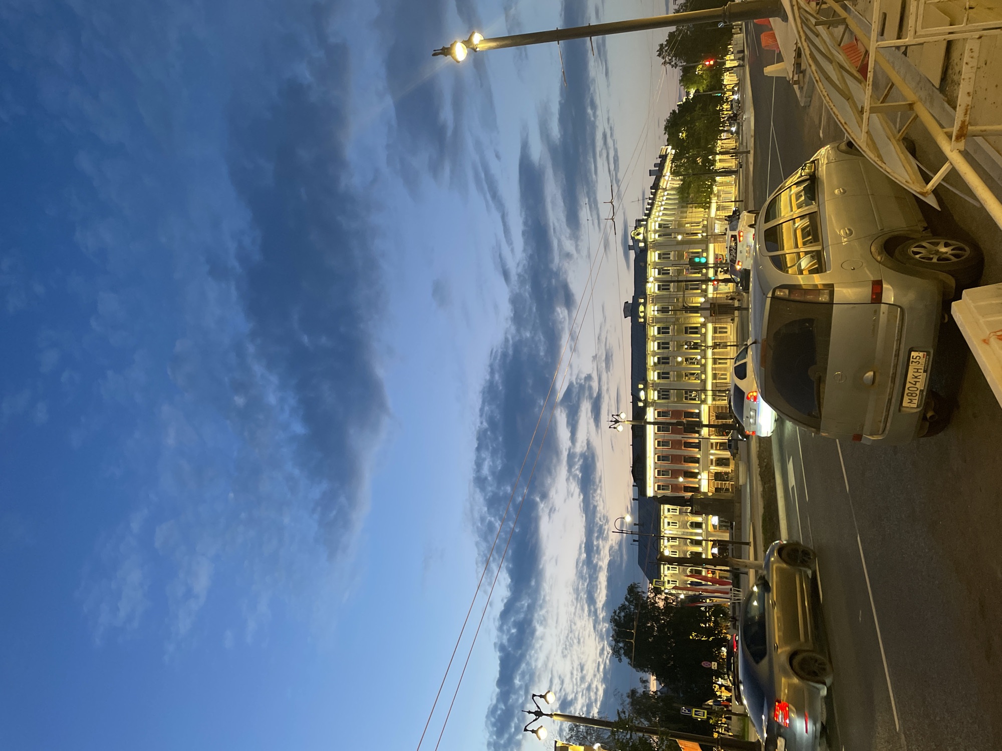 This image captures a twilight street scene in Vologda, Russia, likely on Sovetsky Prospekt, near the intersection with Gorky Street. The sky is a deep blue with scattered clouds, transitioning to a lighter hue near the horizon. A row of illuminated buildings, characterized by their ornate architecture and yellow lighting, lines the right side of the street. In the foreground, several cars are visible, including a silver sedan in the left lane and a silver minivan in the right lane, with its license plate reading "M804KH 35". Traffic lights are illuminated green and red at the intersection. Streetlights with glowing bulbs dot the roadside, and overhead wires crisscross the sky. Trees line the sidewalks, and construction barriers with orange cones are present on the right. The overall impression is of a bustling urban environment at dusk.