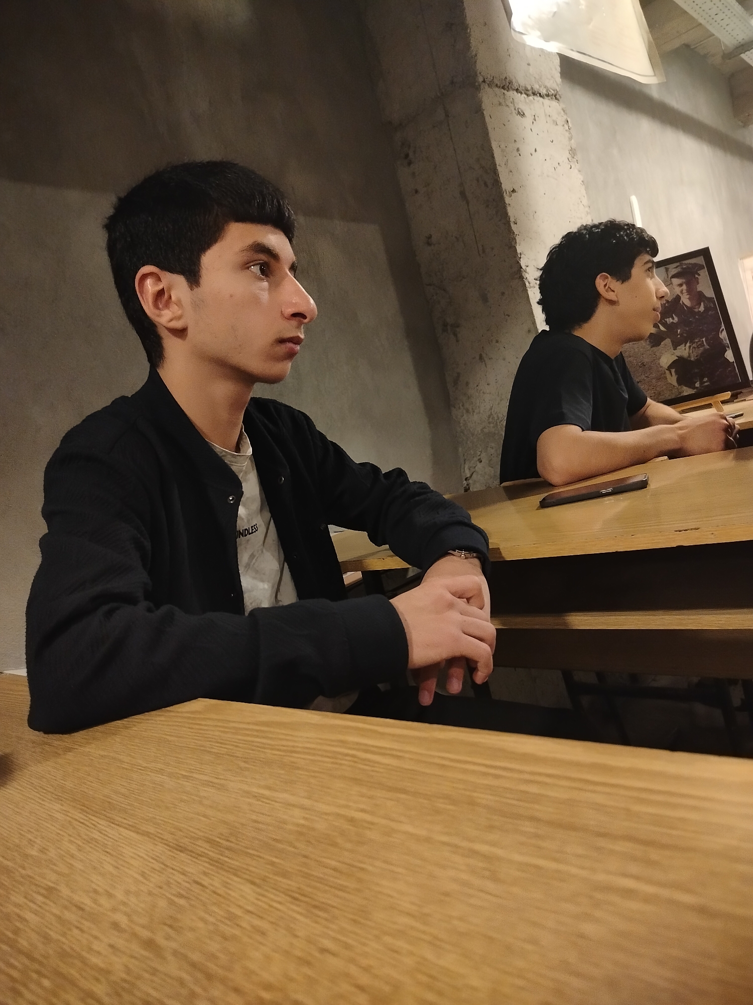 Two young men are seated at a wooden table in what appears to be a cafe or restaurant. The man in the foreground is wearing a black jacket over a white t-shirt, and he has dark hair and a focused expression, with his hands resting on the table. Behind him and to the right, the other man is wearing a black t-shirt and also appears to be focused on something in front of him. A framed picture is visible behind the second man. A cell phone is placed near the second man. The location appears to be in Yerevan, Armenia, based on architectural details and ambient conditions. The setting seems informal, possibly a casual dining or studying area.