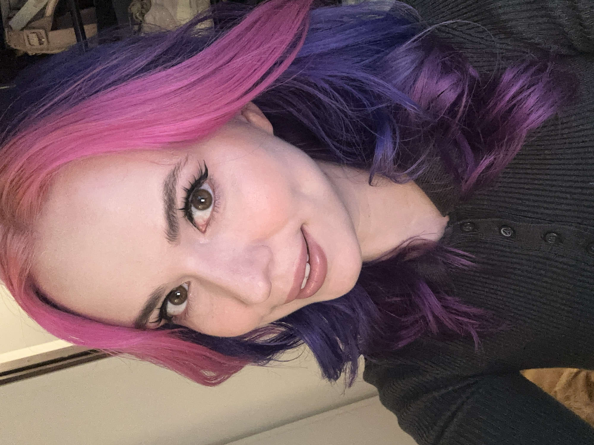 A woman with pink and purple hair, wearing a dark colored shirt with buttons, is looking at the camera and smiling. The background is blurry and indistinct, but a wall is visible. The location is not discernible from the image.