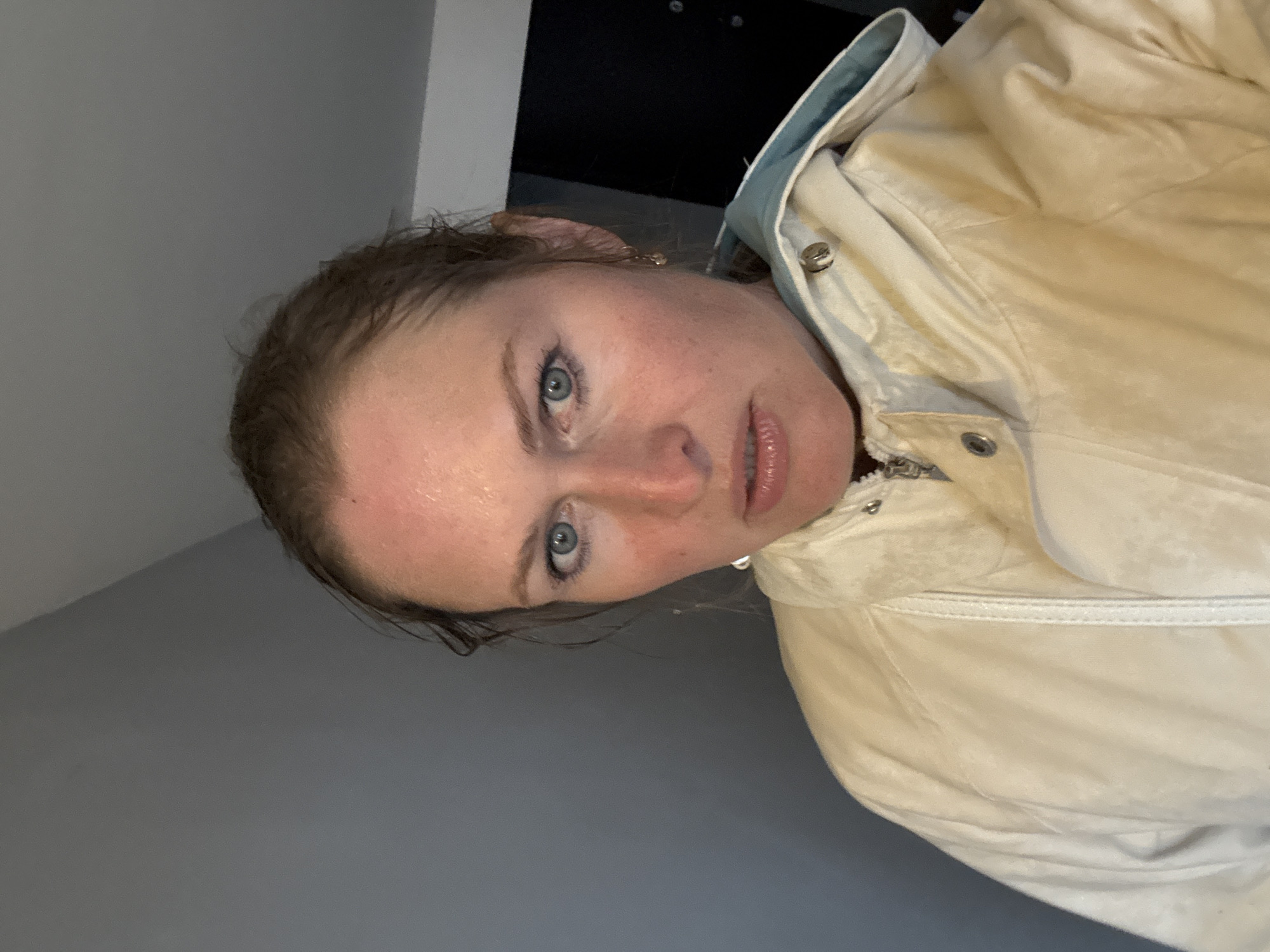 A woman with fair skin and blue eyes is facing the camera. She has wet hair. She is wearing a light-colored jacket with a hood. The background consists of a gray wall and a dark area, possibly a doorway or a piece of furniture.
There is no evidence in the image to suggest the location is Chaikovsky, Russia.