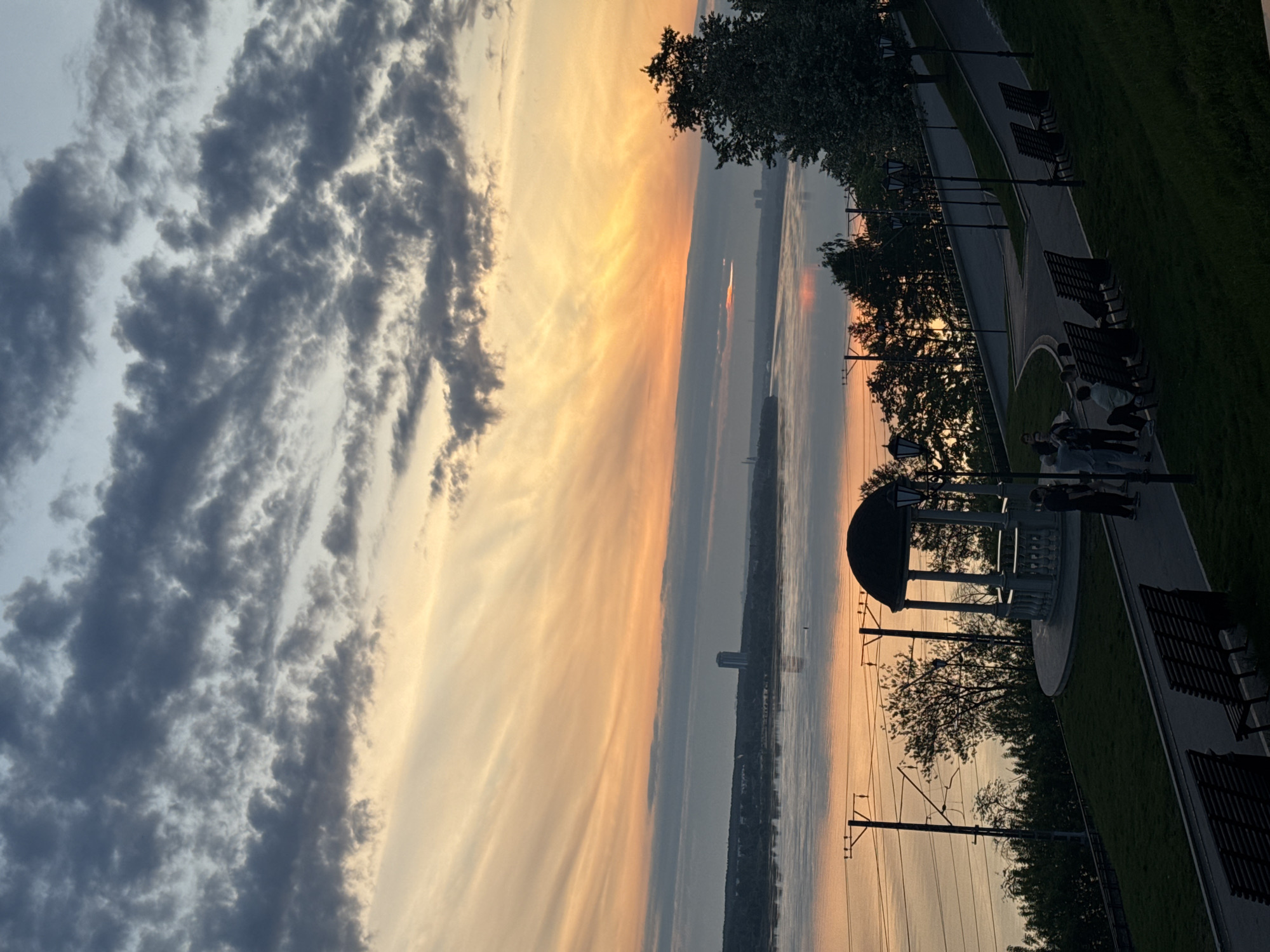 The image depicts a sunset over the Kama River in Perm, Russia. The sky is filled with dark, layered clouds on the left, contrasting with streaks of orange and yellow hues on the right where the sun is setting. The river reflects the colors of the sky. A park embankment runs along the right side of the river, featuring a paved path, benches, and a gazebo structure. People are seen standing near the gazebo. Utility poles with overhead wires are visible along the embankment. A few buildings are visible on the opposite shore of the river.
