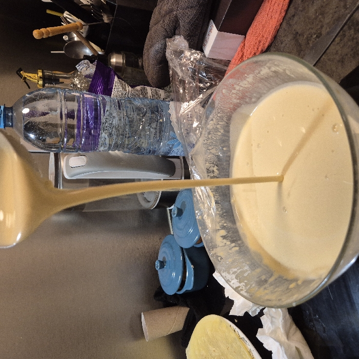 A ladle pours batter into a clear glass bowl. The bowl sits near a blue lidded pot, an empty cardboard tube, and white tissue paper. A water bottle, bag of chips, and cooking equipment reside in the background. An orange towel and gray oven mitt sit behind the bowl. The surface is a dark grey.