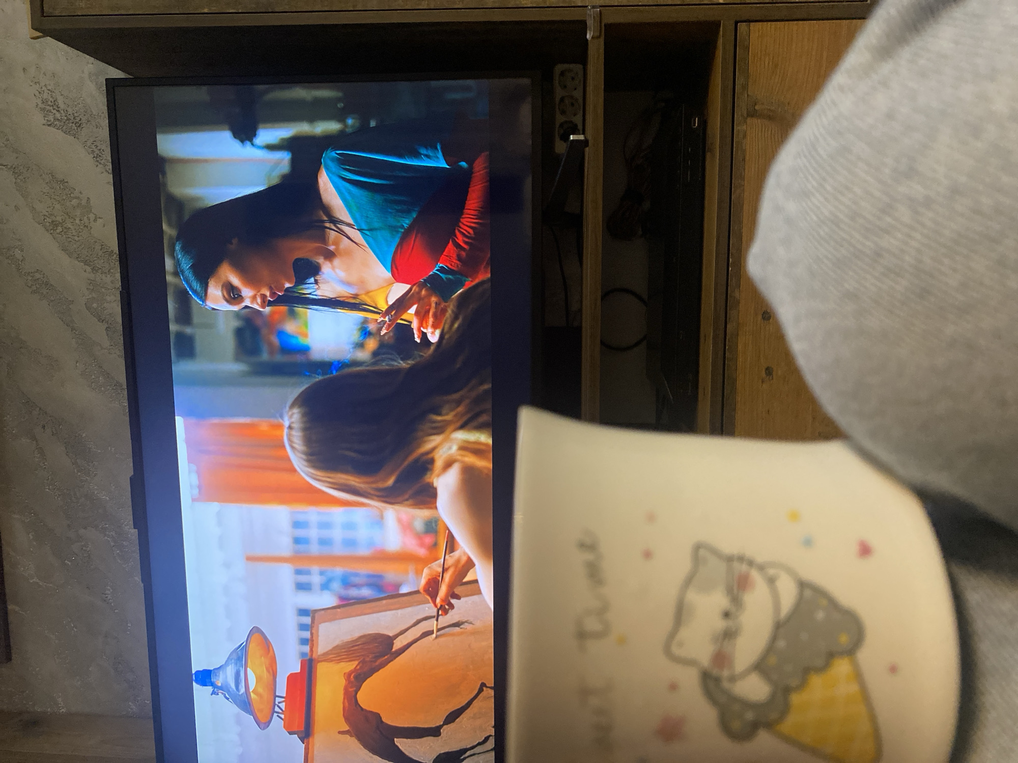 The image captures a person's point of view, watching television in an indoor setting. The main subject, partially visible on the right, is wearing a light grey long-sleeved garment, possibly a hoodie or sweater. In their hand, they hold a white mug with a cartoon depiction of a grey and white cat in an ice cream cone, surrounded by small colorful shapes. Faint text, likely "Sweet Time," is visible on the mug.
The television screen, mounted within a wooden shelving unit, displays a scene vertically. On the screen, two female figures are visible. The upper figure has dark hair and is wearing a teal top with red sleeves, appearing to be cutting something with scissors. The lower figure, with long brown hair, is engrossed in painting on a canvas. The painting depicts a stylized, stretched human-like figure with flowing hair.
To the left of the TV, a textured grey wall is visible. Above the TV and to its right, part of a wooden cabinet or shelving unit is seen, housing a power strip with multiple outlets and various electronic cables and devices. The lighting suggests an indoor setting, likely in the evening or at night. The scene on the TV appears to be from a show or movie.