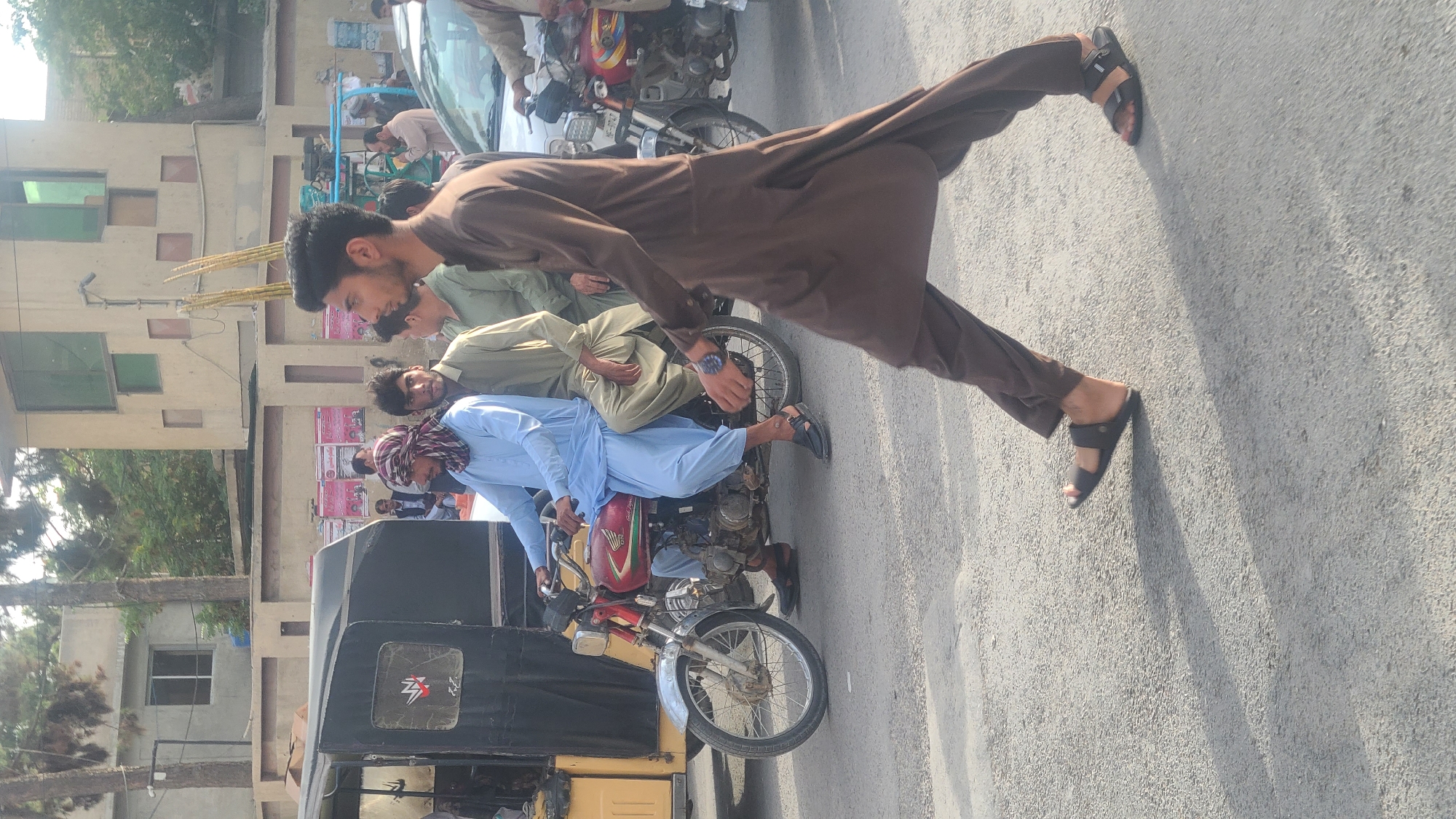 This is a medium shot of a street scene in Quetta City Tehsil, Pakistan. In the foreground, a man in a brown shalwar kameez and sandals walks from right to left, partially obscuring a motorcycle with two men on it. The man driving the motorcycle wears a blue shalwar kameez and a patterned headscarf. The passenger behind him wears a light green shalwar kameez. Behind them, a black auto-rickshaw is visible. Further in the background, other individuals and vehicles are present, including a white car and another motorcycle. The environment appears to be an urban setting with buildings and trees lining the street. The lighting suggests it is daytime.