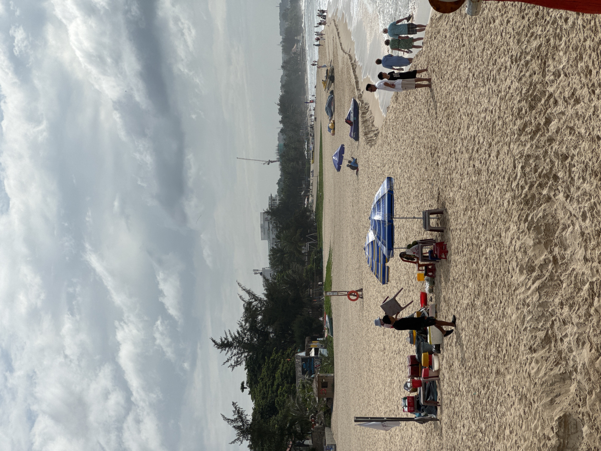 The image depicts a sandy beach with the ocean and a cloudy sky in the background. The location is identified as Mũi Né, Vietnam. In the foreground, a man is carrying a chair across the sand. To his left, there are several red buckets, some yellow containers, and a white cooler stacked on a wooden stand. Two blue and white striped umbrellas are set up on the beach, one with a small table and chairs underneath. Further down the beach, a group of people, including a man, a child, and three women, are walking towards the water. Several jet skis and other watercraft are visible on the sand near the water's edge. In the distance, there are buildings, palm trees, and a construction crane. The overall scene suggests a day at the beach with recreational activities and vendors.