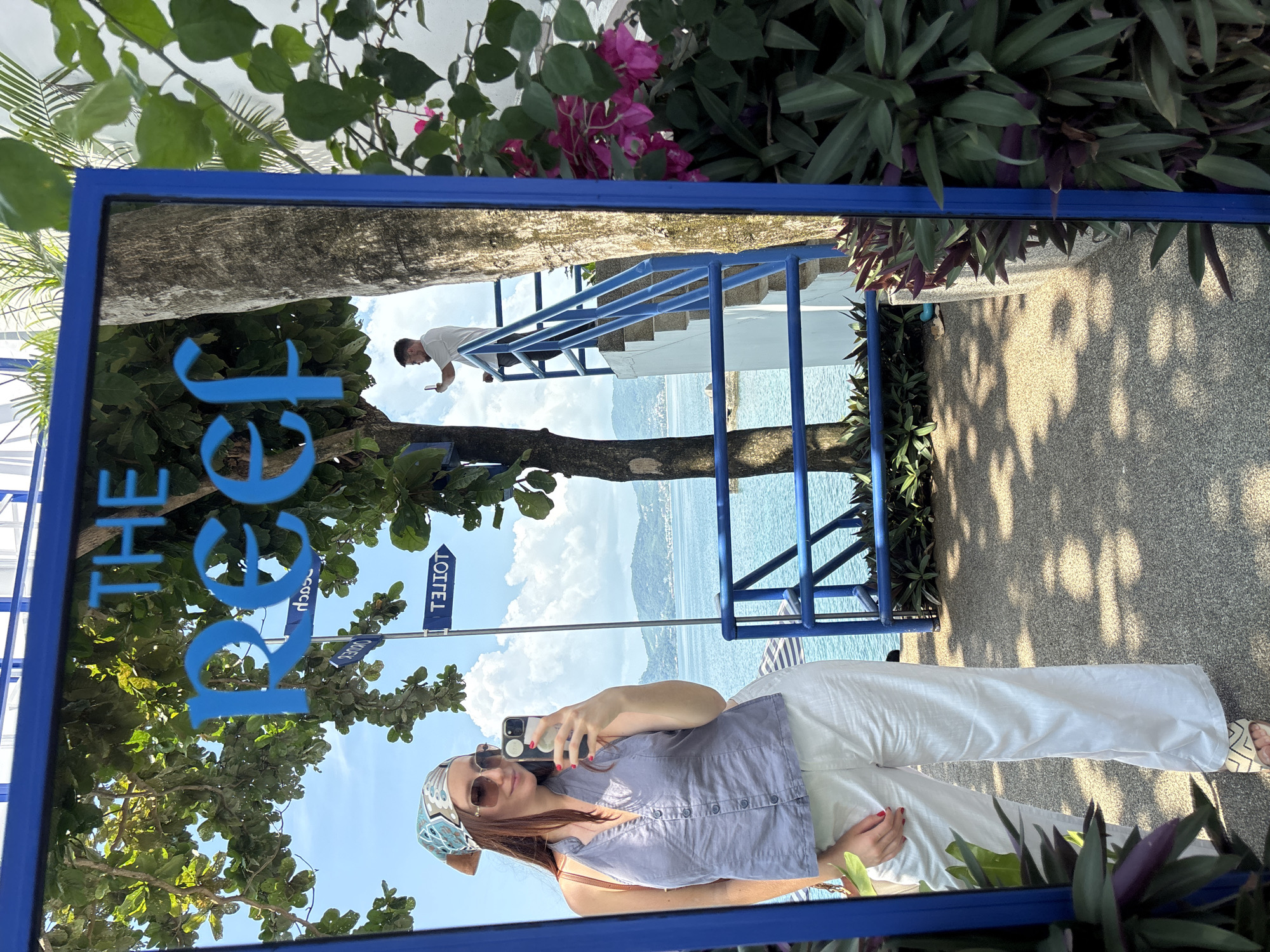 A selfie is taken in a large mirror with a blue frame, showing "The Reef" sign in blue. The woman in the selfie is wearing a gray button-down shirt, white pants, sunglasses, and a patterned headscarf. She is holding a smartphone in her right hand. The mirror reflects a scene with a man on blue stairs, a tree, and a view of a body of water, with foliage visible around the mirror's edge. A directional sign indicates "Hotel" and "Beach." The image suggests a location in Kamala, Thailand, given the coastal setting and tropical foliage.