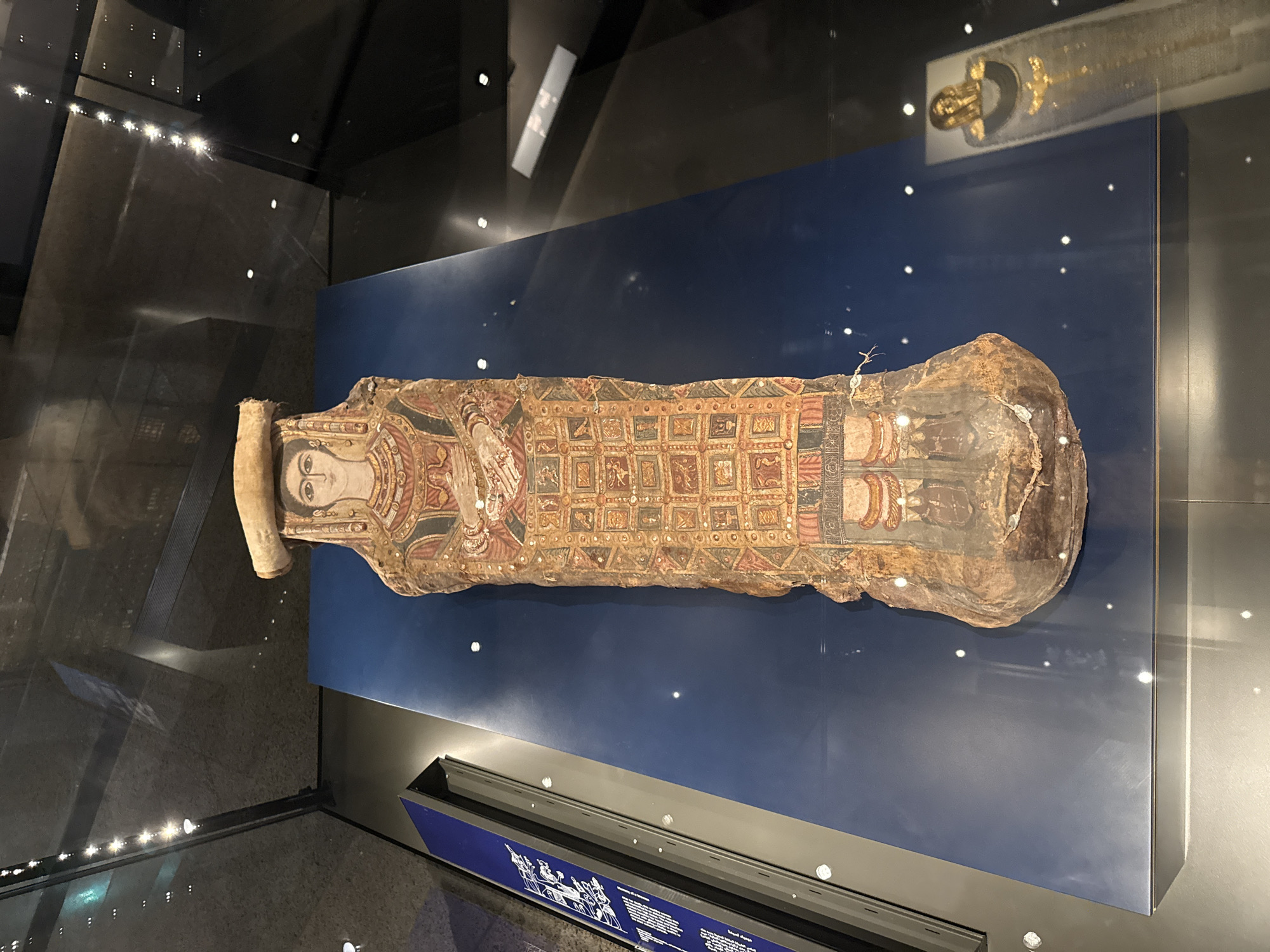 A mummified sarcophagus stands upright on a dark blue display platform within a museum exhibit. The sarcophagus is intricately painted with depictions of a human face, arms crossed over the chest, and a long, patterned robe adorned with geometric shapes and figures. The head of the sarcophagus is topped with a light-colored headdress. To the left, a blue exhibit panel with text and images is partially visible. In the background, a second sarcophagus, this one appearing to be covered in gold, is reflected in the glass enclosure. The lighting in the exhibit is characterized by several bright spots and reflections on the glass. The overall scene suggests an archaeological or historical display, likely in a museum.