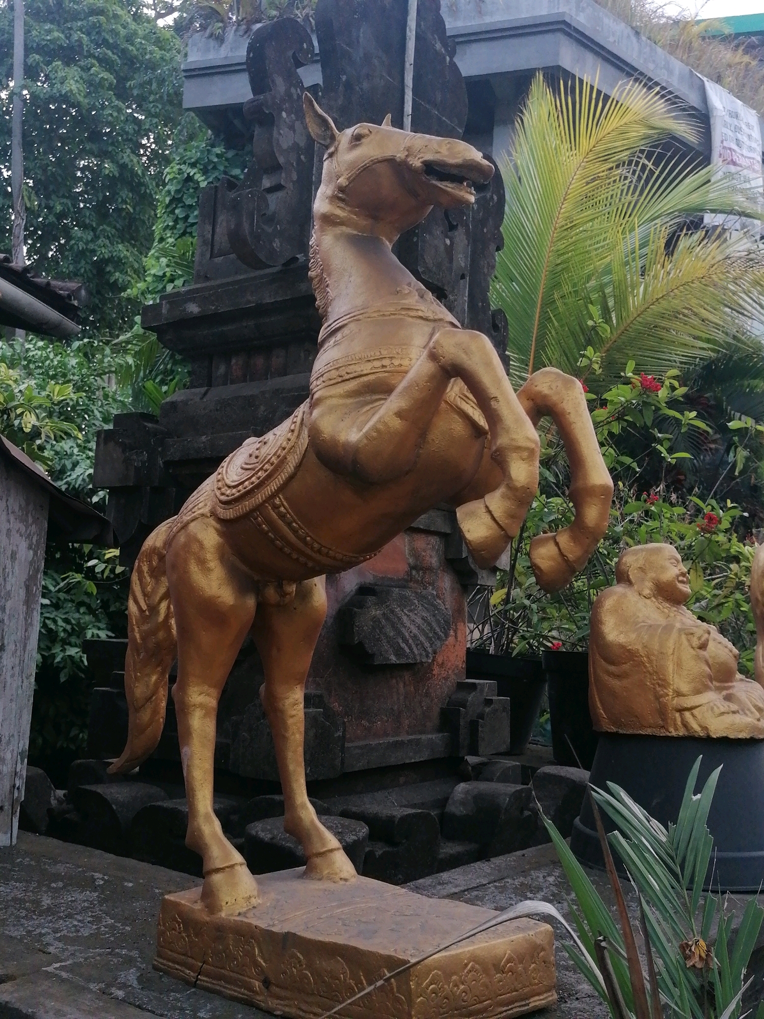 A golden statue of a horse rearing on its hind legs stands prominently in the foreground of this outdoor scene. The statue is detailed, with a saddle and ornate bridle. To the right, a smaller golden statue of a seated figure, possibly a Buddha or a deity, is visible. Behind the horse statue, a dark stone structure, likely an entrance or monument, is partially obscured by lush green foliage. A palm tree with yellowing fronds arches over the scene, suggesting a tropical or subtropical environment. The ground appears to be paved with stone or concrete. The overall impression is one of an outdoor decorative element in what could be a temple or garden setting in Ubud District, Indonesia, characterized by traditional Balinese artistry and natural elements.