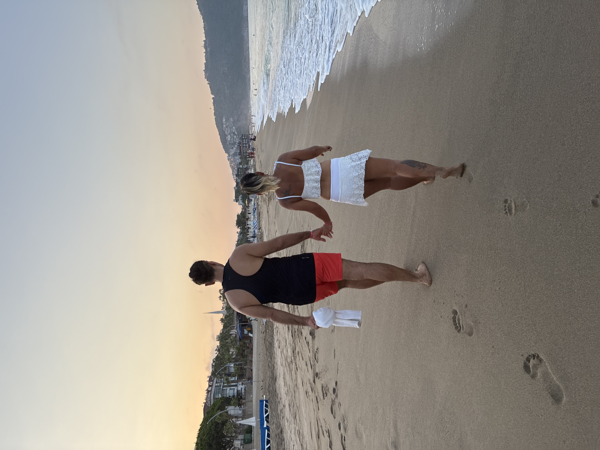 A couple walks hand-in-hand along a sandy beach in Alanya, Turkey at sunset. The man, wearing a black tank top and red shorts, carries a white towel. The woman, in a white bikini set, walks beside him. The ocean is on their right, with gentle waves lapping the shore. Buildings and hills can be seen in the background. Footprints mark the wet sand where the water has receded. The sky displays a soft gradient of orange and blue, characteristic of twilight.