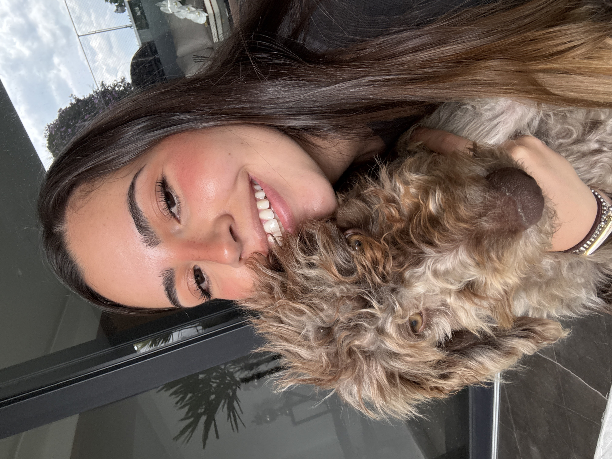 A person is holding a dog and smiling at the camera. The person has long brown hair, is wearing a dark top, and is wearing a bracelet on her left wrist. The dog is brown and tan with curly fur. The background includes a reflective surface, likely a window, with a fence and cloudy sky visible in the reflection. The exact location in San Bernardino Tlaxcalancingo, Mexico is not identifiable based on the visual information provided.