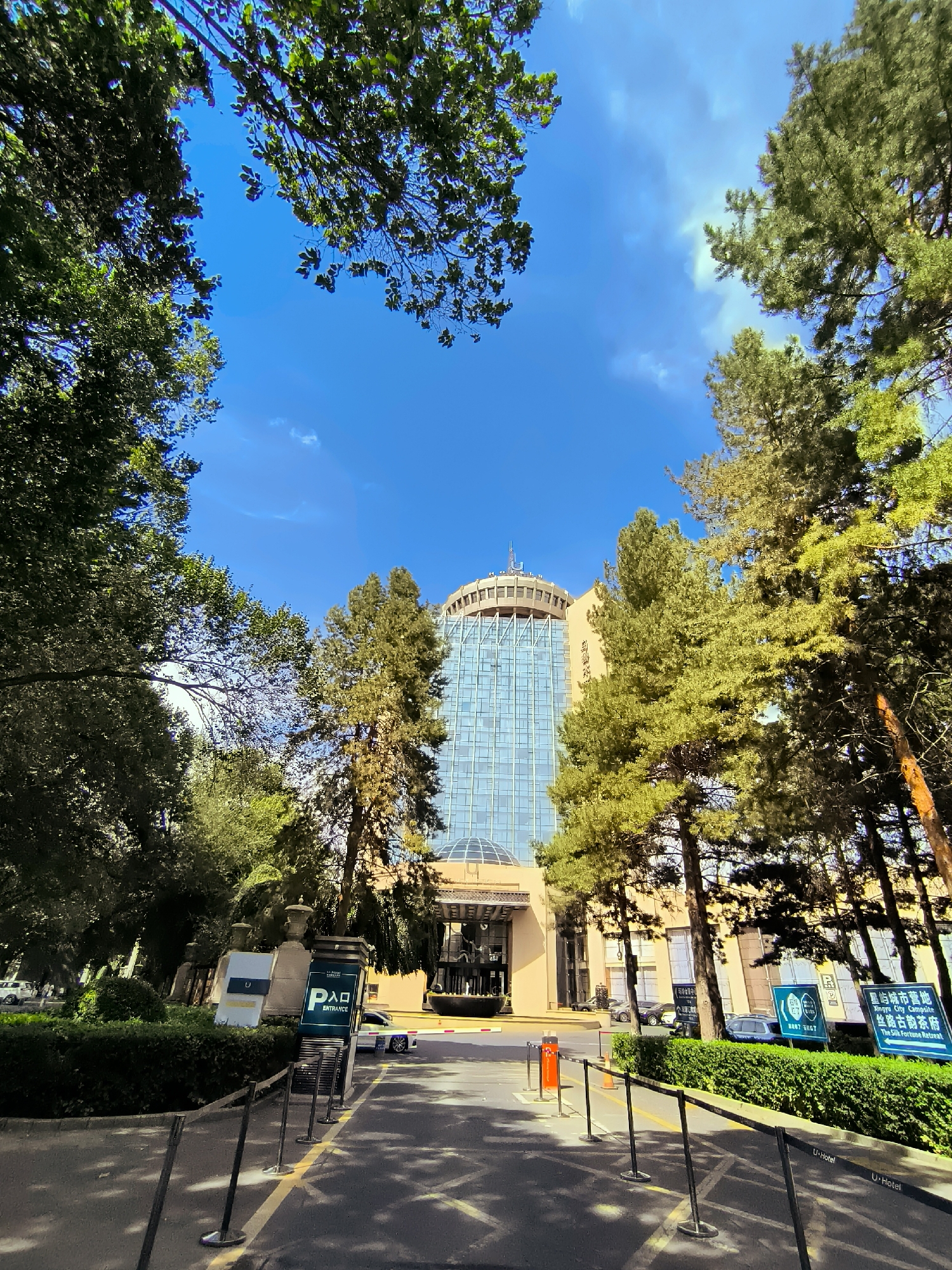The image shows the entrance to the U-Hotel in Ürümqi, China. The hotel is a modern building with a glass facade, surrounded by trees. The entrance is marked by a sign with the hotel's name and logo, and a barrier that appears to be for a vehicle entrance. The sky is clear and blue. The scene appears to be set in a well-maintained urban environment.
