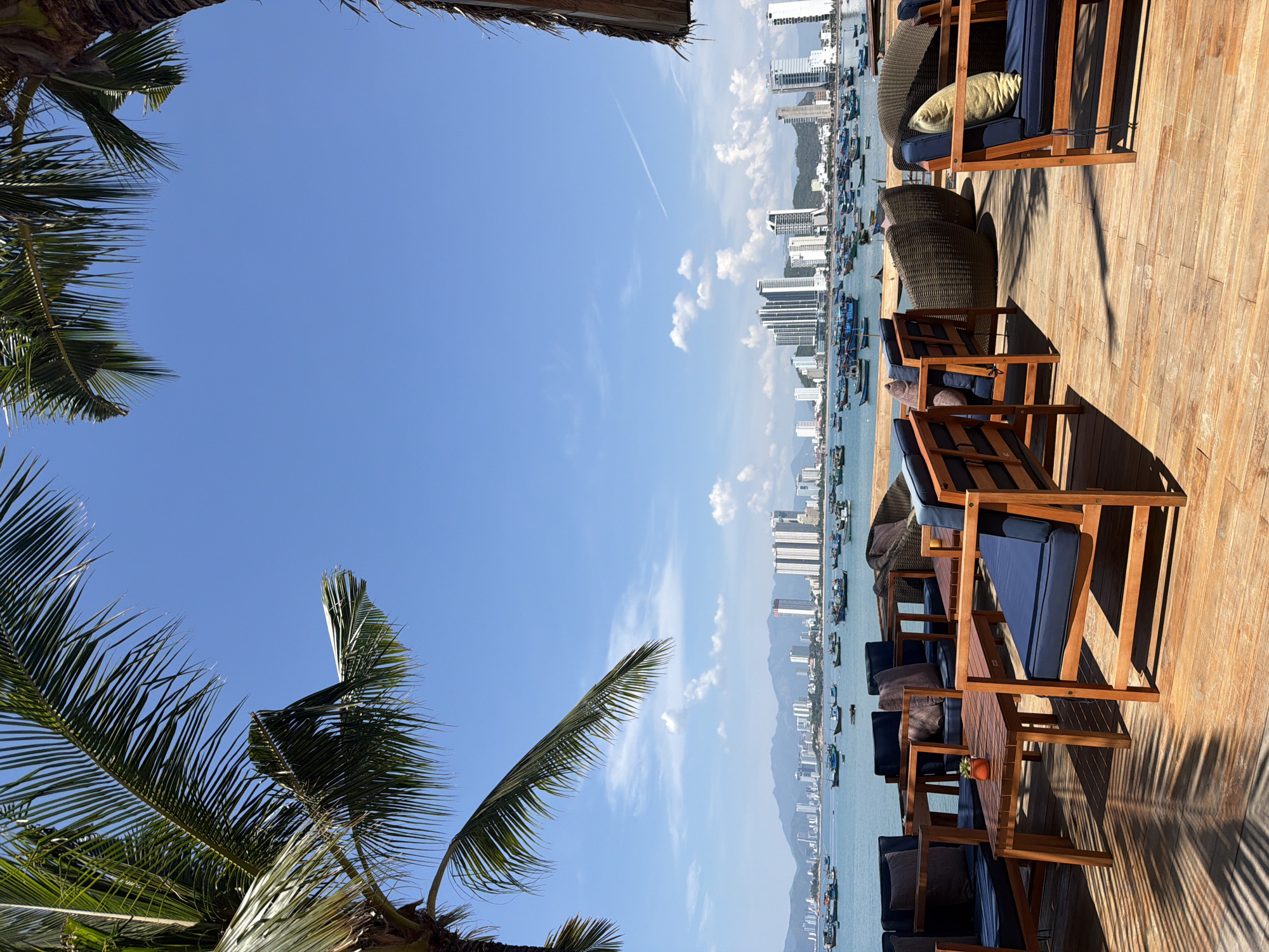 A brightly lit, outdoor scene shows a wooden deck area with multiple seating arrangements. The chairs have wooden frames and blue cushions. Additional seating includes woven, rounded chairs. The deck overlooks a body of water, with buildings and mountains visible in the distance, indicating a coastal city. Numerous boats dot the water. Palm trees frame the left side of the image. The city is Bac Nha Trang Ward, Vietnam.