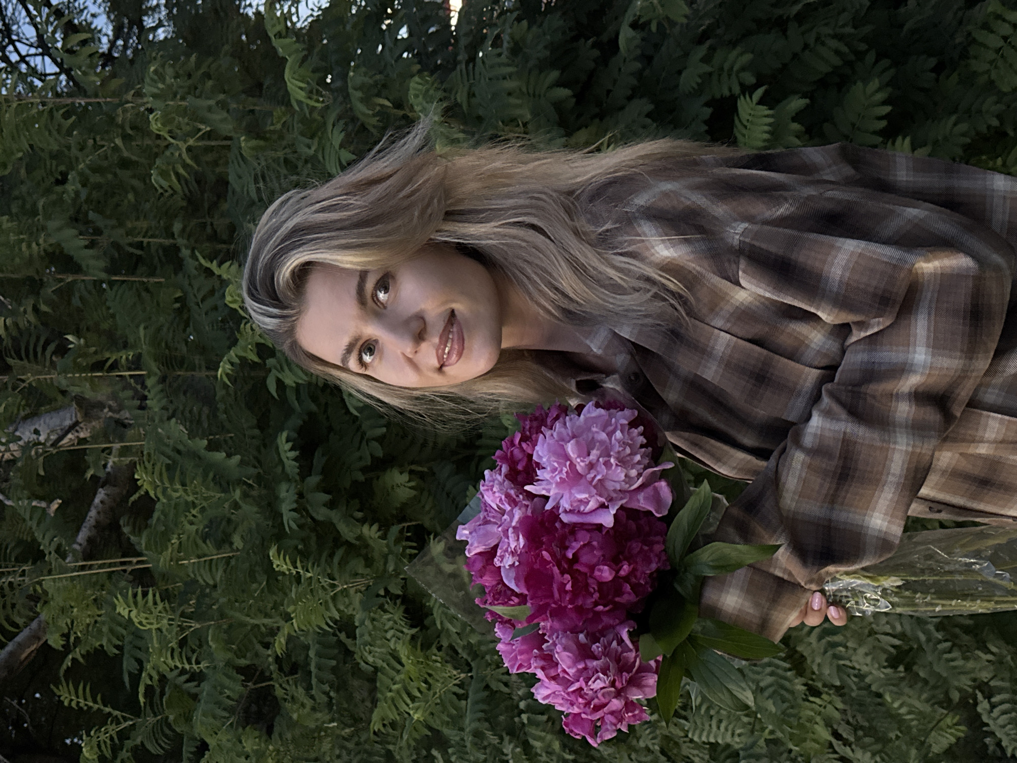 A medium-shot, waist-up portrait of a fair-skinned woman with long, wavy blonde hair is featured against a backdrop of lush green foliage. She is looking towards the viewer with a subtle smile, her head slightly tilted. The woman is holding a bouquet of vibrant pink and deep magenta peonies with green leaves in her left arm, which is partially obscured by the flowers and her garment. She is wearing a brown and beige plaid button-up shirt. The lighting suggests it is either early morning or late evening, with a soft, diffused light illuminating the scene. The background consists of dense, leafy trees, creating a natural and serene environment. The overall tone of the image is pleasant and calm.