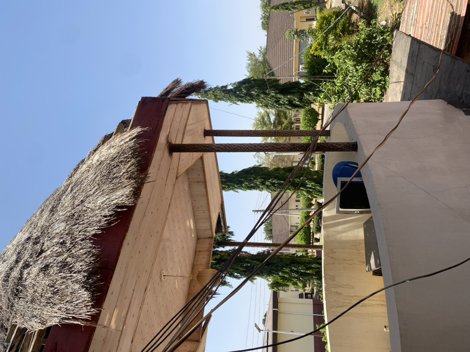 The image captures an exterior scene under a clear blue sky in Tudun Nupawa, Nigeria, viewed from an elevated vantage point. In the immediate foreground, a curved, light-colored concrete parapet wall with visible surface cracks partially encloses an area. Within this enclosure, a dark-colored rectangular object and a blue plastic chair are discernible. Multiple black cables descend from the upper left, crossing the frame diagonally and resting on the parapet wall.
Above the parapet, a structure features a prominent thatched roof composed of dry straw, supported by a dark red fascia board and a light-colored, paneled underside. Two textured brown columns support this roof structure. A single, light-colored pendant fixture hangs from the paneled underside.
Beyond the immediate structure, the environment includes multiple tall, slender green trees and various green foliage. In the middle distance, several single-story buildings with light-colored walls and brown-tiled roofs are visible, interspersed among the greenery. A solar-powered street light and utility poles with overhead wires are present on the left side of the background. No individuals are visible within the frame.