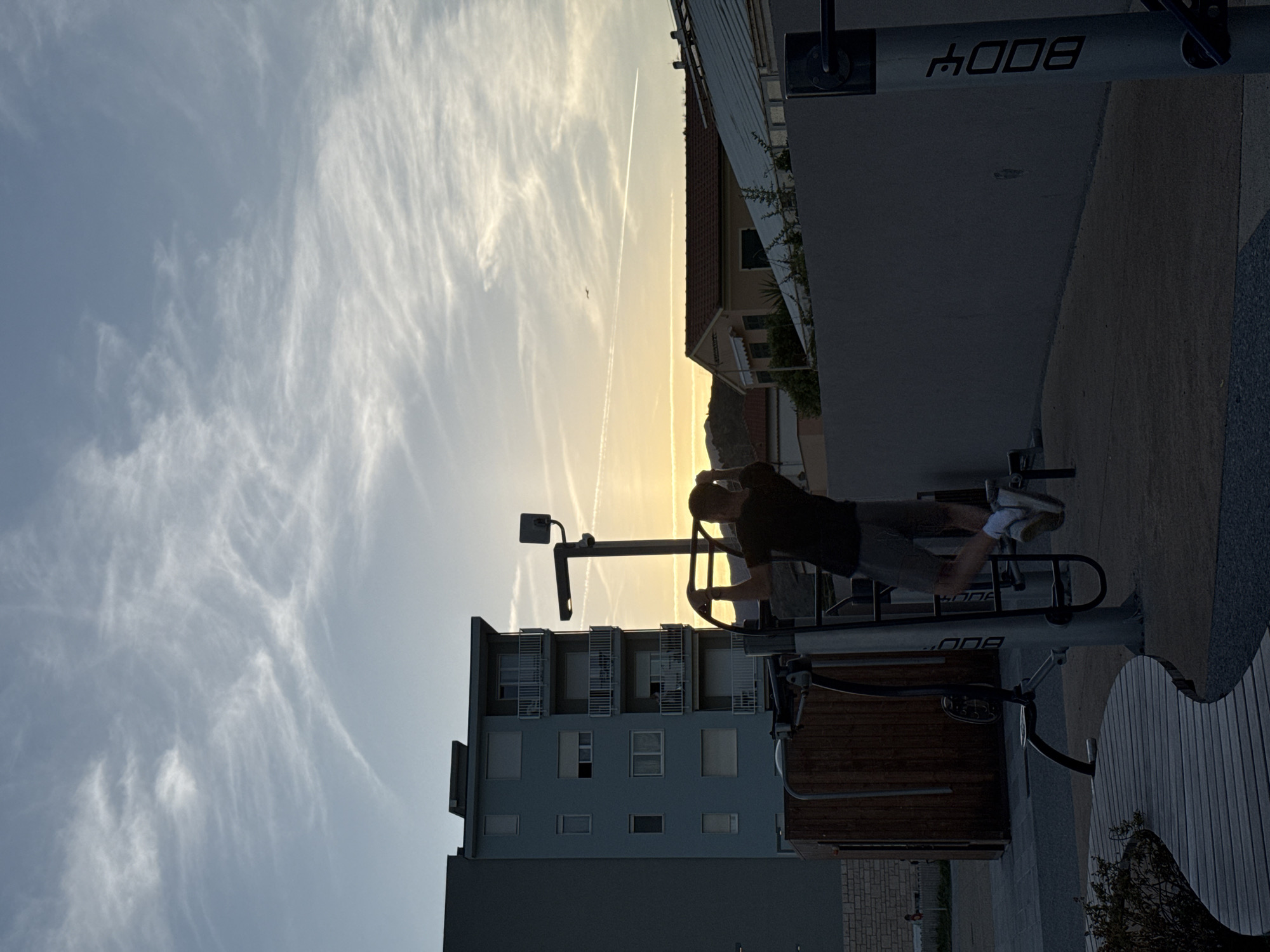 A man is performing pull-ups on an outdoor gym apparatus at sunset in Vallecrosia al Mare, Italy. The man, silhouetted against the sky, wears a dark t-shirt and shorts. The gym equipment is positioned in front of a tall, light-blue apartment building. In the background, other buildings with tiled roofs are visible, and the sky is filled with wispy clouds and contrails, indicating the setting sun. The ground is paved with gravel and features a curved wooden seating area. A small airplane is visible in the distance.