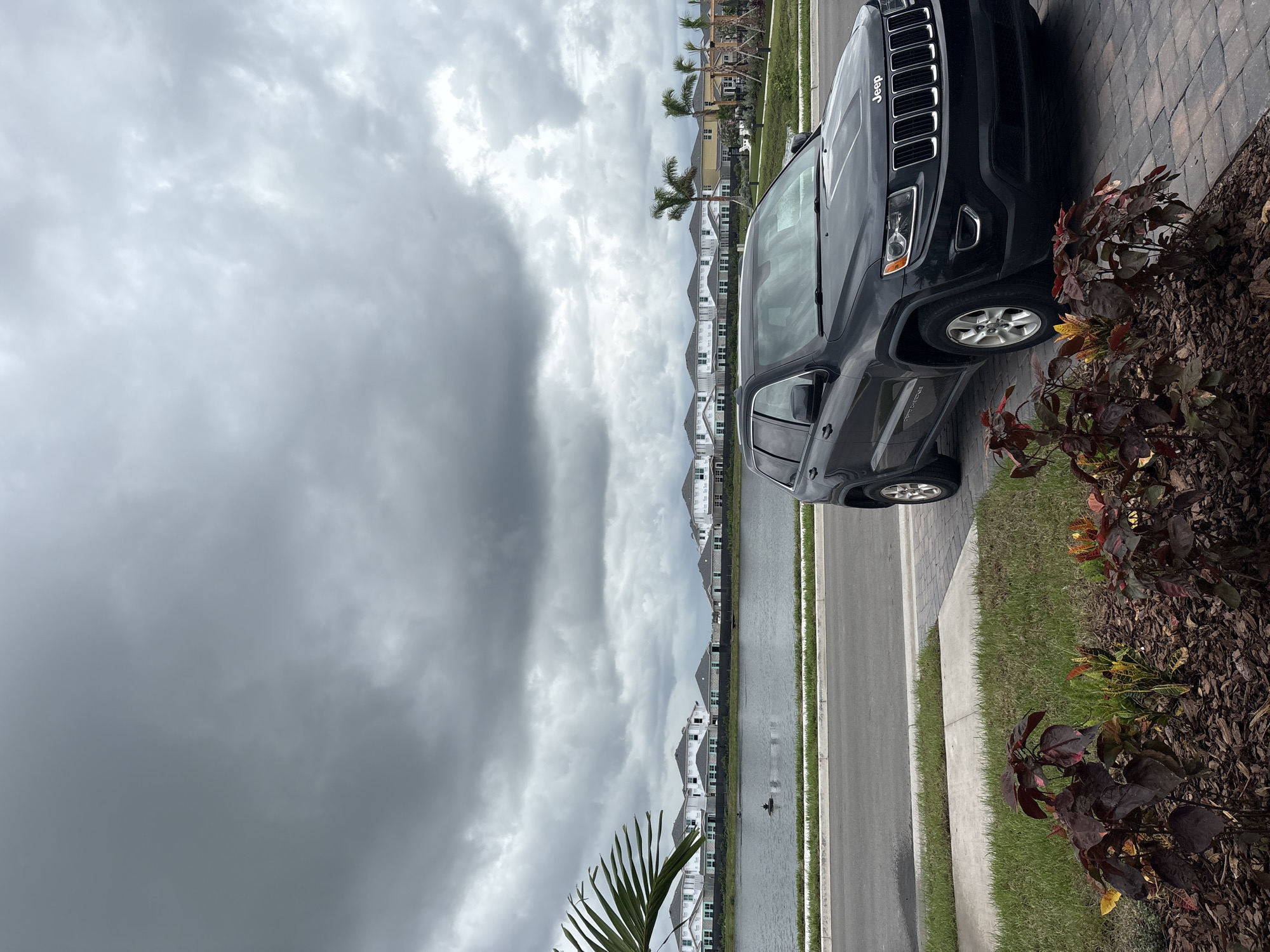 An overcast day in Lakewood Ranch, United States. A dark-colored Jeep Grand Cherokee is parked in a driveway, partially obscuring a manicured lawn and a paved walkway. In the background, a calm lake separates the foreground from a row of newly constructed houses with white exteriors and dark roofs. Tall palm trees are visible along the lake's edge. The sky is filled with heavy, dark gray clouds, suggesting impending rain.