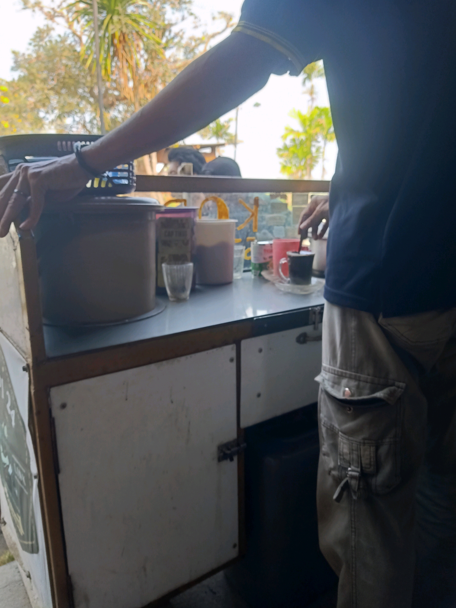 A person, seen from the chest down, stands at a food cart in Kalipecabean, Indonesia. They wear a dark blue t-shirt and tan cargo pants, with their left hand resting on a black basket on the cart. The cart's countertop is cluttered with various items, including large containers, a small glass, and cups. In the background, lush greenery and palm trees suggest an outdoor setting, possibly a street or park. The overall lighting indicates it is daytime.