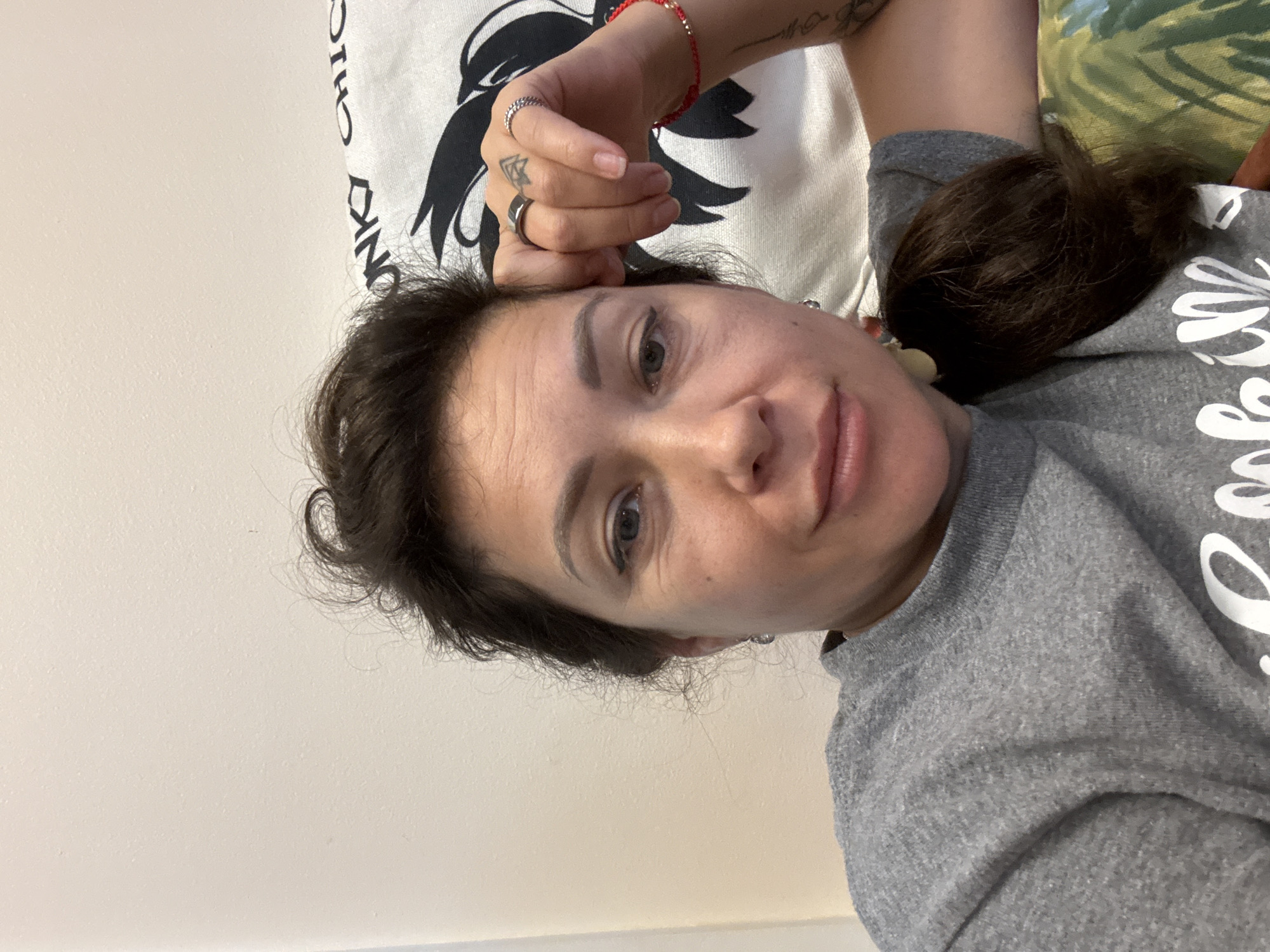 A medium shot selfie is taken of a woman with brown hair and fair skin. She is wearing a grey sweatshirt and has her right hand resting on her forehead. She is looking at the camera with a slight smile. Behind her, a white pillow with a black graphic is visible, as well as part of a green and white patterned couch. The image is taken indoors, and the lighting suggests it is daytime. No specific location in Key West is discernible from the image.