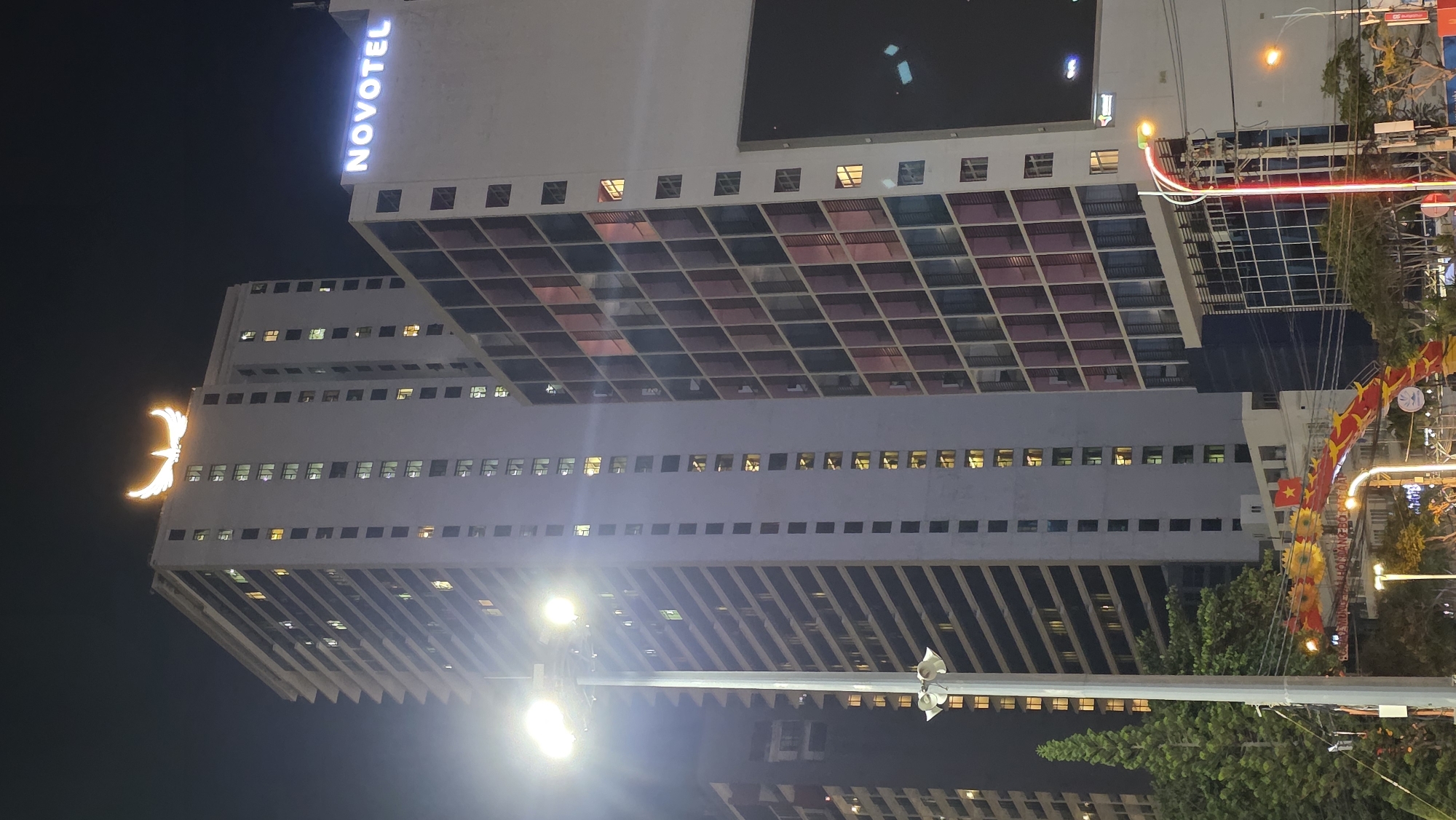 The image presents a vibrant city night scene dominated by tall, illuminated buildings. On the left, a prominent modern skyscraper features the illuminated "NOVOTEL" sign vertically displayed near its peak. This building has a distinctive architectural design with a grid of square windows in varying shades, and numerous smaller windows along its lower sections, many of which are lit, indicating activity within.
To the right of the Novotel, another tall building stands, adorned with a large, bright golden glowing symbol resembling a stylized phoenix or bird with outstretched wings, positioned on one of its upper corners. This building also displays rows of illuminated windows.
At street level, in the lower right portion of the image, the area is brightly lit by multiple artificial sources, including prominent street lamps. Green trees are visible, and the scene is festive with decorations. A long, red and yellow garland or banner, possibly a stylized dragon or festive string lights, stretches across the street. A small Vietnamese flag is also discernible among these festive elements.
The time of day is clearly night, as indicated by the dark sky and the reliance on artificial lights. The presence of the Vietnamese flag and the type of festive decorations strongly suggest a location in Vietnam, specifically Nha Trang as per context, likely during a celebratory period. The visible text is "NOVOTEL" on the hotel building.