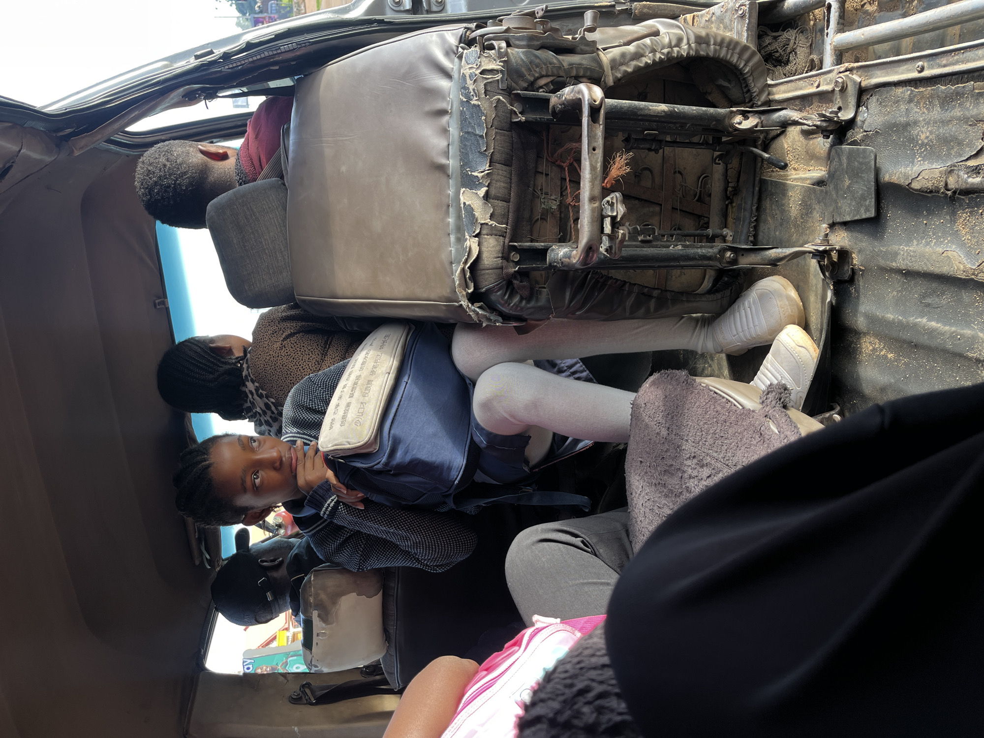 In Gura Top, Nigeria, a group of people are traveling inside a crowded vehicle, possibly a public transport van. The interior of the vehicle appears worn and aged, with damaged upholstery on the seats and a dirty floor covered in sand.
In the foreground, a young girl with braided hair is seated, looking to her right with a thoughtful expression. She is wearing a grey patterned sweater and white stockings. She is holding a blue backpack on her lap, which has a cream-colored pouch attached to it. Next to her, a person wearing a pink top is visible, with their arm resting on their lap. Further back, another person wearing a black baseball cap is seated. Behind the girl, two more individuals are seated, one wearing a patterned top and the other a maroon shirt. The windows of the vehicle are open, revealing a bright, sunny exterior with hints of buildings and trees in the distance. The overall scene suggests a typical day of travel in the region, highlighting the resourcefulness and daily routines of its inhabitants.