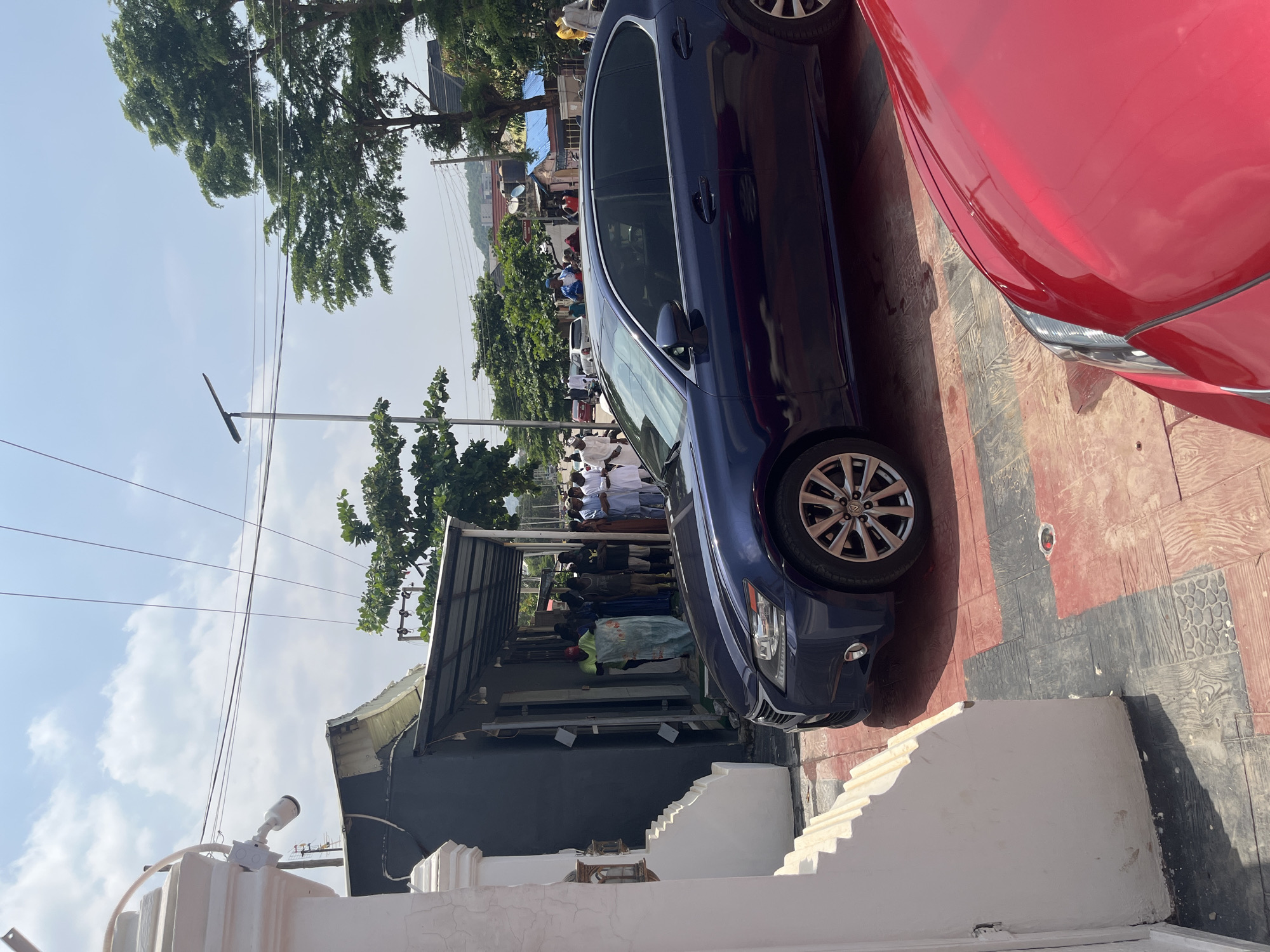 The image depicts an outdoor scene on a sunny day, likely in Abuja, Nigeria. The focus is on a dark blue car parked on a patterned tiled surface. To the right, a portion of a red car is visible.
In the background, a street scene unfolds. Several people, some wearing white traditional attire, are gathered near a low-rise building with a metal roof. The building appears to be a shop or stall, with items displayed outside, suggesting a market or commercial area. Trees with lush green foliage line the street, and electrical poles with wires are visible against a bright blue sky with scattered white clouds.
A CCTV camera is mounted on a white structure in the foreground, indicating a potentially monitored area. The overall atmosphere seems to be that of a bustling daytime environment, possibly in a populated urban or semi-urban setting. No specific event is clearly identifiable, but the gathering of people suggests social interaction or commerce. The lighting indicates it is daytime, likely morning or afternoon given the brightness of the sun and the clear sky.