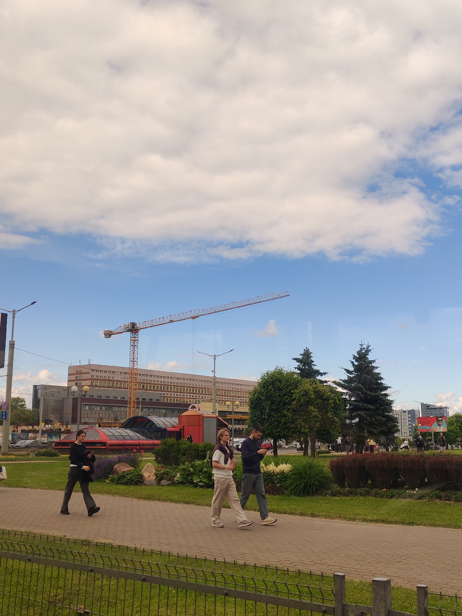 In an outdoor urban scene, three individuals are walking along a paved path. One person, wearing a black sweater and dark pants, walks with their head down, looking at their phone. Another person, dressed in a white t-shirt and light-colored pants, walks with their hands in their pockets, a sweater tied around their neck. The third person, in a dark blue sweater and grey pants, is looking at their phone while walking.
The setting appears to be a park or public walkway in front of a modern building. Behind the walkers, a large, multi-story building with a distinctive reddish-orange, arched structure in front of it is visible. Further in the background, a tall construction crane stands against a bright blue sky with scattered white clouds. Trees, including some evergreen trees, and landscaped bushes are also present. The time of day appears to be daytime, given the lighting. The overall mood seems casual, with people engaged in everyday activities. No text is clearly discernible in the image.