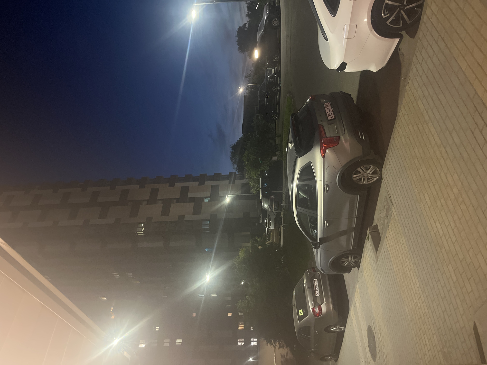 The image depicts an urban street scene at night in Khimki, Russia, illuminated by artificial light sources. The deep blue sky above indicates either dusk or night conditions.
In the left background, a tall, multi-story residential building stands prominently. Its facade features a modern, geometric pattern composed of alternating dark and lighter rectangular panels. Numerous windows are visible across its surface, with many displaying illuminated interiors, suggesting occupancy. A powerful, unidentifiable light source positioned near the upper left of the building generates a bright starburst lens flare.
The foreground consists of a patterned pavement made of light-colored rectangular paving stones, with a drain cover visible on the ground. Adjacent to this pavement is an asphalt road.
Three primary vehicles are visible in the foreground and mid-ground:
1. On the far left, parked parallel to the pavement, is a light beige Skoda Rapid sedan, facing away from the camera. It bears a Russian license plate **O120HC 799**. A yellow sticker is affixed to its rear window.
2. In the center-right, parked partially on the paved area and partially on the road, is a grey Cadillac XT4 SUV, also facing away. Its Russian license plate reads **K363HT 977**. The vehicle features roof rails and dual exhaust tips.
3. On the far right, only the rear right quarter of a white Lada Vesta sedan is visible, parked on the paved area. It displays a distinctive wheel design.
In the background, along the road, several other vehicles are present. A dark Hyundai Palisade SUV is parked, followed by another white SUV. Further down the road, a dark sedan with illuminated headlights is visible.
Multiple pole-mounted streetlights are active throughout the scene. A prominent streetlight on the right side of the image casts a strong glow and creates a large lens flare, while another is visible further down the road in the background. Dense green trees and shrubs are situated between the paved areas and the road, and also in the distant background. No people are visible in the scene.