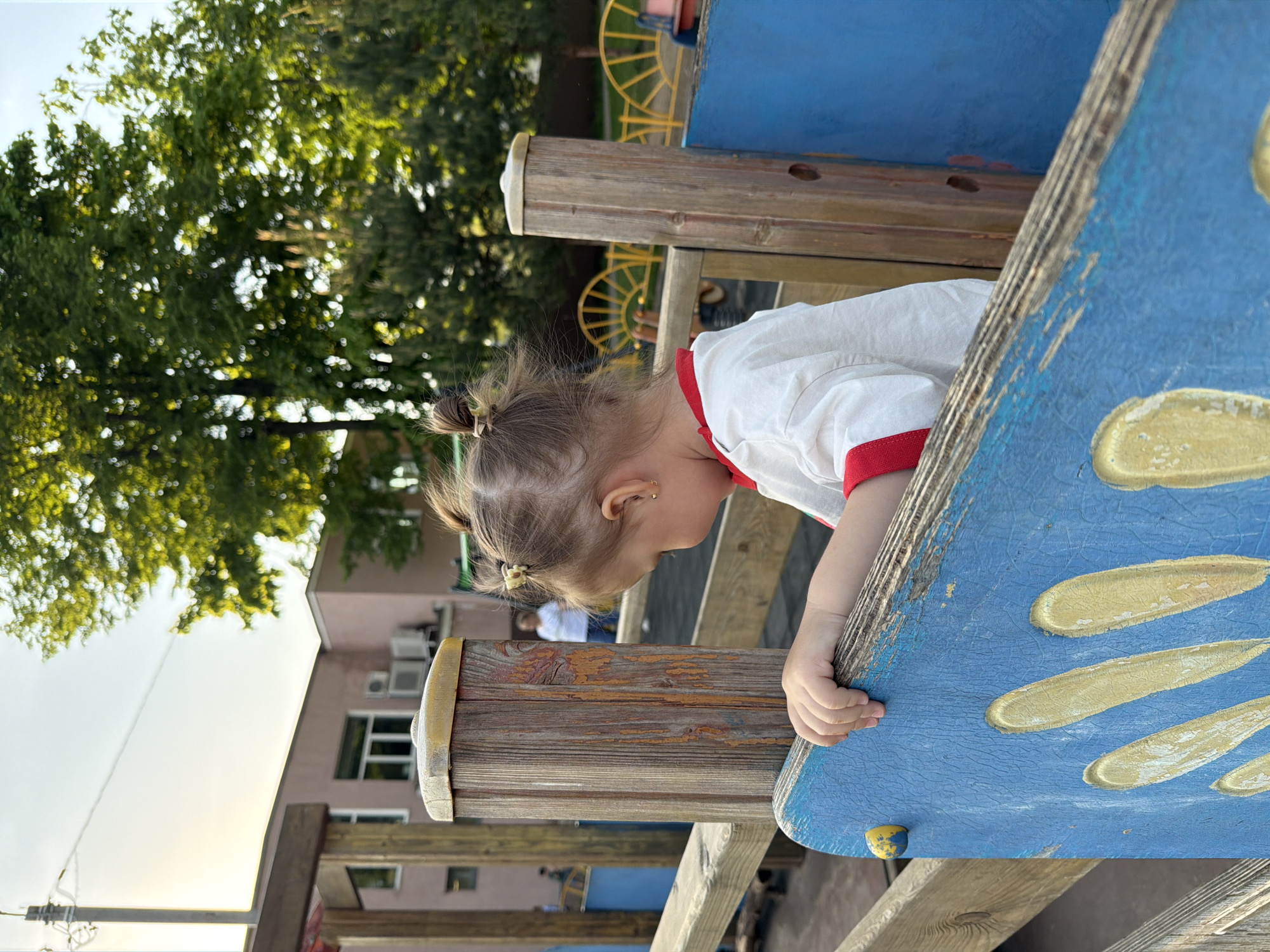 A toddler is captured in an eye-level shot on a playground in Reshetnikovo, Russia. The child, with a small ponytail held by a white hair tie, wears a white shirt trimmed with red around the collar and sleeves. The toddler is bent over, hands gripping the edge of a blue wooden structure that appears to be part of the playground equipment. The blue surface has yellow oval designs.
The playground is surrounded by greenery and a building with windows and air conditioning units visible in the background. The lighting suggests it is daytime. A wooden fence with yellow elements is also partially visible.