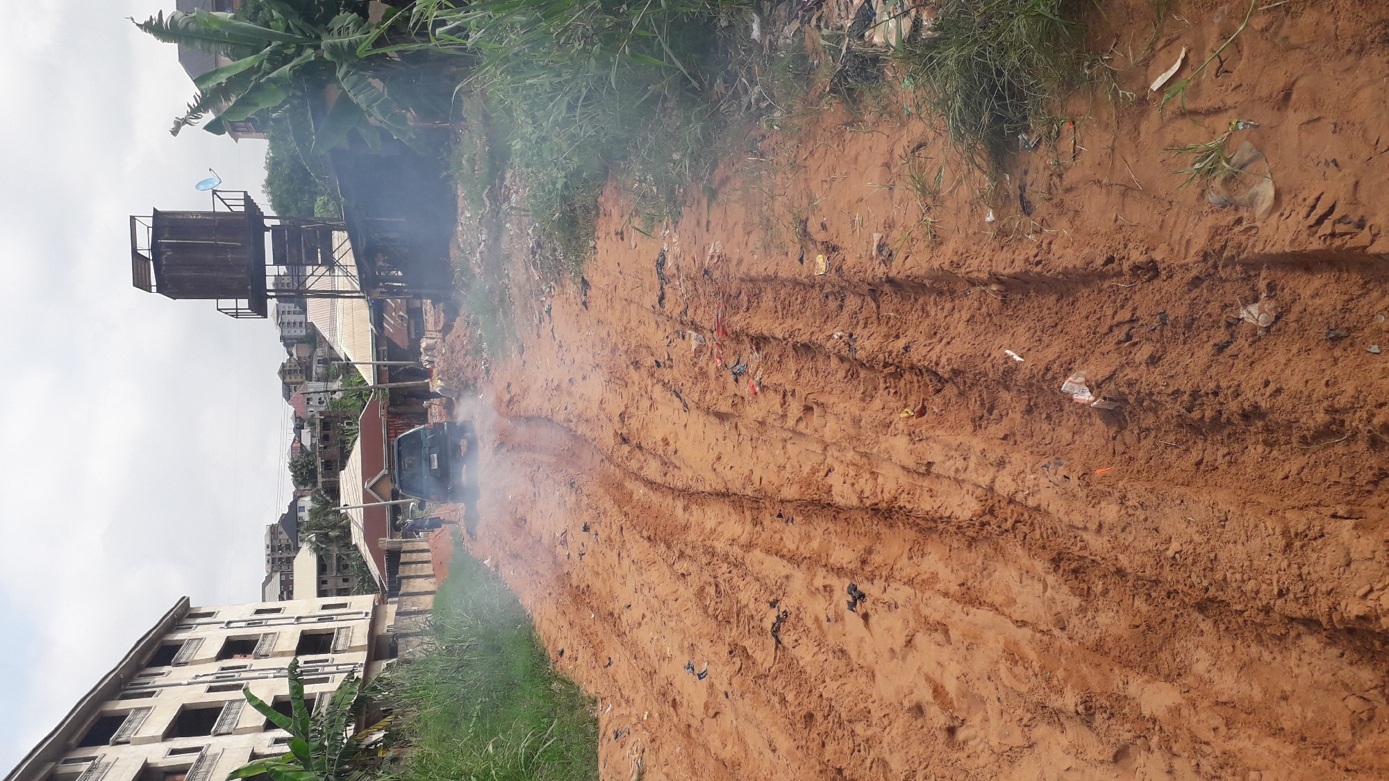 The image depicts a rural road in Nkpor, Nigeria, characterized by a reddish-brown dirt surface with prominent tire tracks. The road is littered with various forms of debris, including plastic bags, paper, and other discarded items. In the middle ground, a dark-colored SUV is visible, emitting a plume of white exhaust smoke as it drives away from the camera. To the left of the road, a multi-story building, possibly under construction, stands tall, with lush green vegetation in the foreground. To the right, a rustic water tower with a metal tank on a raised platform dominates the skyline, accompanied by more vegetation and debris. The overall environment suggests a developing or unpaved area with signs of both construction and neglect. The sky is overcast, indicating a cloudy day.
