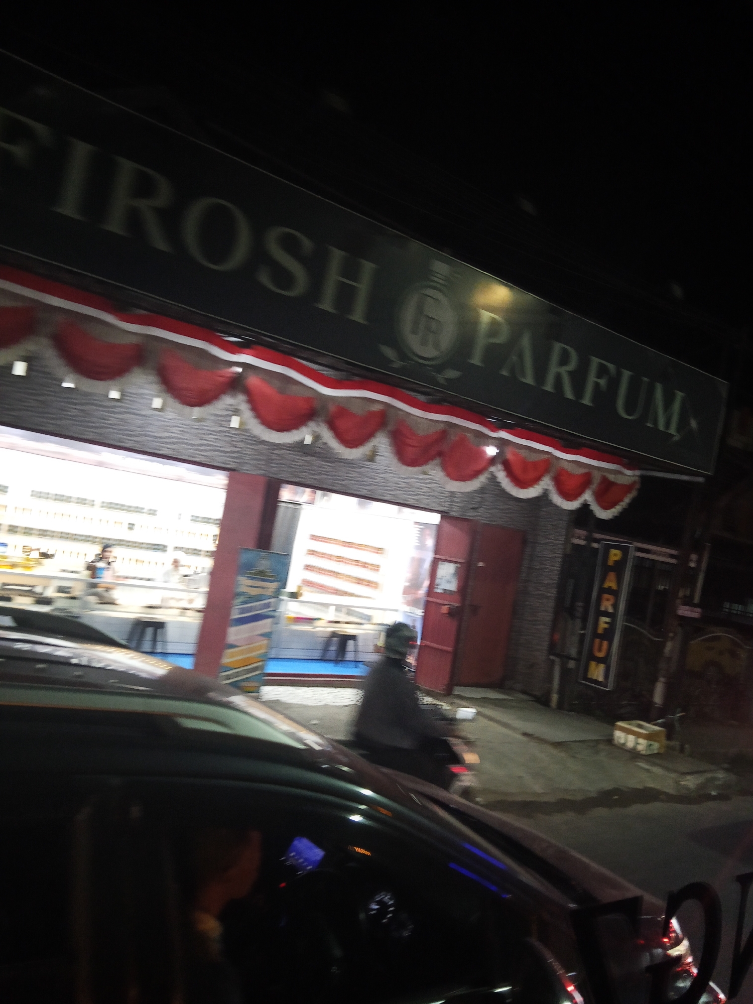 The image depicts a street scene at night, likely in Marumpa, Indonesia, focusing on a store with a prominent sign reading "FIROSH PARFUM". Red and white bunting decorates the store's facade. Inside, a brightly lit interior shows shelves stocked with items, suggesting a retail environment. To the right of the store entrance, a vertical sign also reads "PARFUM". A person is visible riding a motorcycle and another individual is standing near the store entrance. In the foreground, the roof and hood of a dark-colored car are visible, indicating the photo was taken from within a vehicle. The surrounding environment appears to be a street with pavements and buildings.
