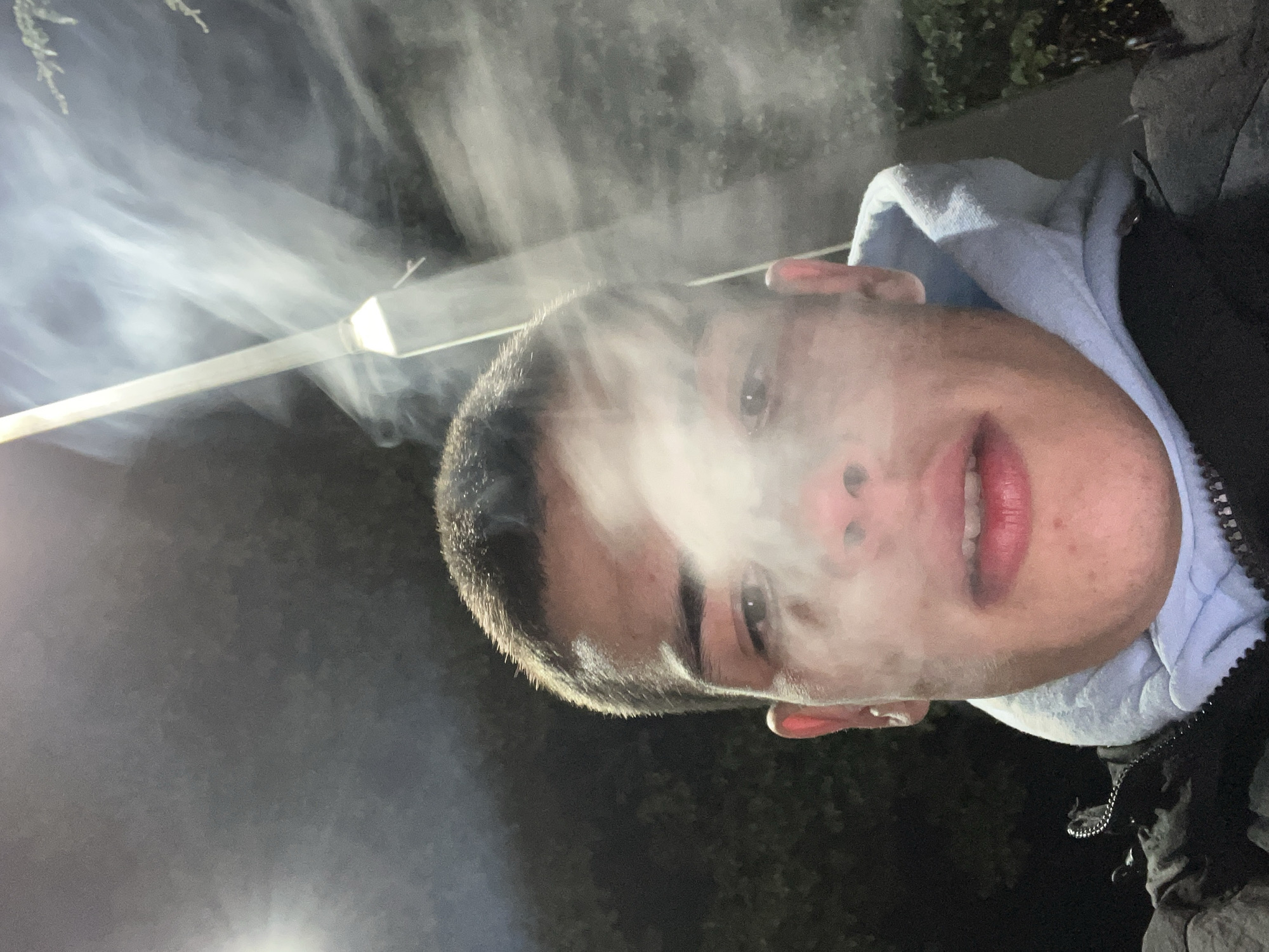 A young man with short, dark hair and light skin is facing the camera. He appears to be exhaling smoke, as wisps of white vapor obscure the area around his nose and mouth. He is wearing a hooded jacket, with the hood a light blue color. The background is dark, with some foliage visible. To the upper left, there is a horizontal light source, possibly artificial. There is no information to suggest the location is Karavelovo, Bulgaria.