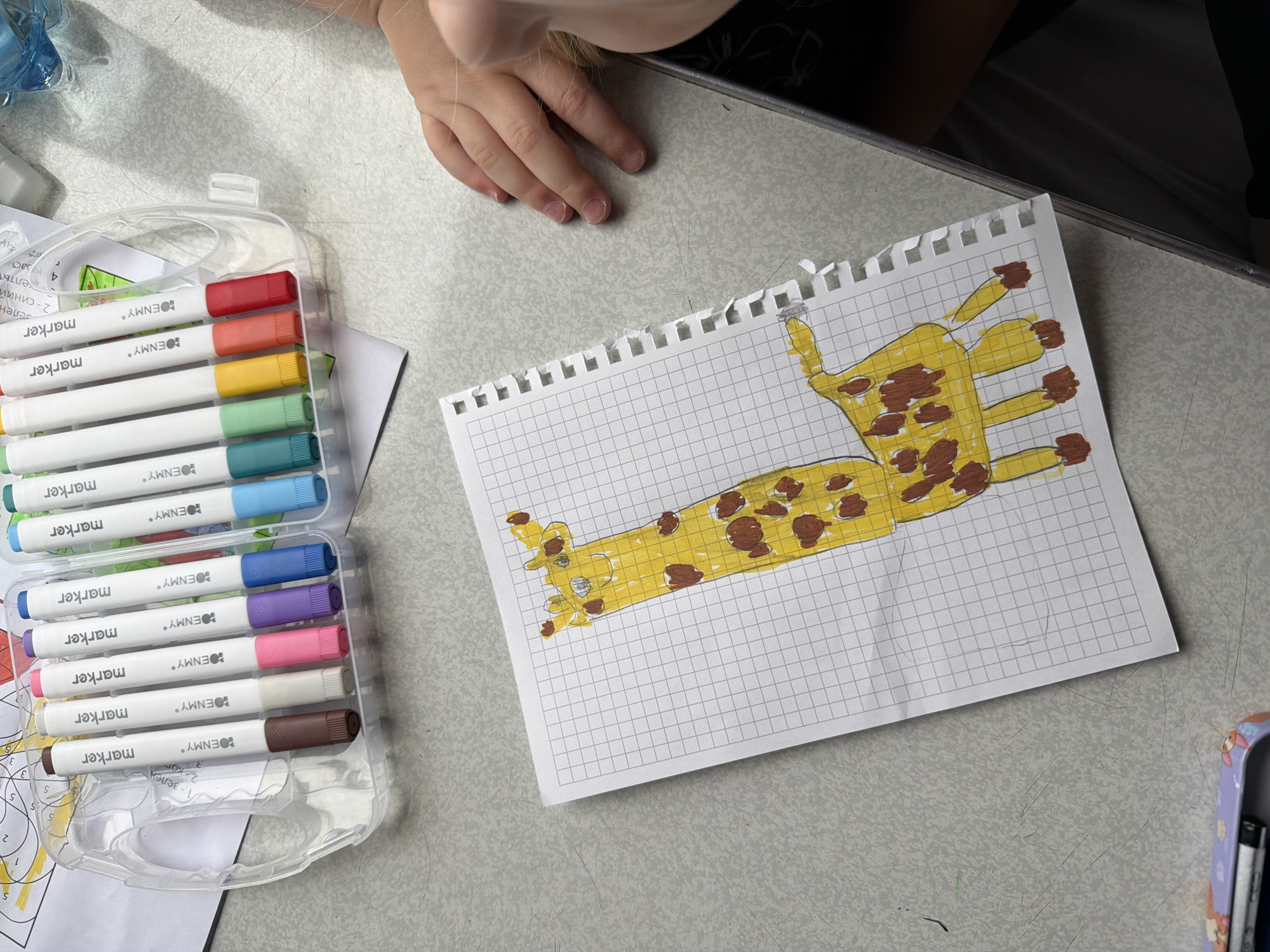 A child is engaged in an art activity, seen from a high-angle perspective, likely at a table or desk. The main subject is a sheet of graph paper with a hand-drawn and colored giraffe, depicted in yellow with brown spots, lying horizontally on the grid lines. Next to the drawing, a clear plastic case holds a set of "BENMY" brand markers in various colors, including red, orange, yellow, light green, blue, dark blue, purple, pink, light grey, and brown.
Part of a child's hands and forearm are visible in the upper left corner of the frame, resting on the light-colored, textured surface of the table. A blue bottle or container is partially visible in the far upper left. Below the marker case, other papers or a coloring book are stacked, showing text in Cyrillic script. Visible words include "2 - синий" (blue), "1-зелен" (green), "келты", and "4-краст" (red), along with the number "5" repeated, indicating a potential color-by-number activity in Russian. The scene suggests a calm, creative atmosphere, likely indoors during daytime. The presence of Russian text aligns with the given location context of Uzunovo, Russia.