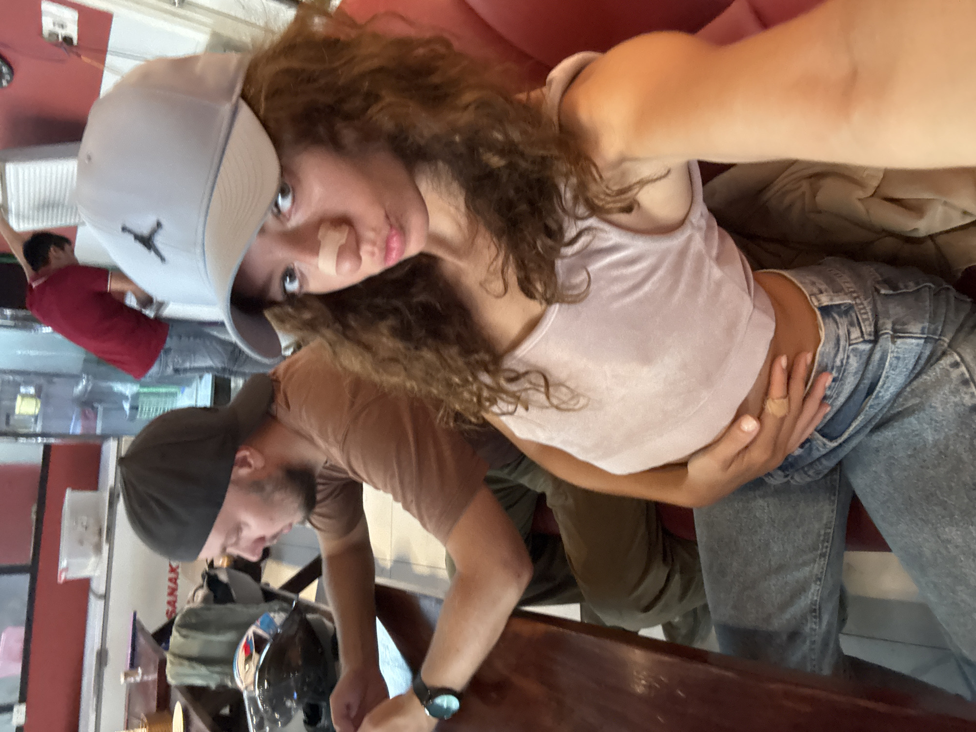 A selfie shows a woman indoors with a man at a table, in what appears to be a café setting. The woman, wearing a light gray baseball cap with a logo and a pale pink tank top, looks directly at the camera with a bandage on her nose. Her hair is curly. She is seated on what looks like a red sofa. Her light wash denim jeans are visible. A man in a brown t-shirt stands at a table in front of her, his back to the camera and head turned to his left, wearing a dark baseball cap. He is looking down at an object on the table. Part of a motorcycle helmet sits on the table. A silver watch is on his left wrist. Behind them, another man in a red shirt stands facing left. The location is likely within the city of Đà Nẵng, Vietnam, as evidenced by the letters "SANAK" visible on a sign in the background.