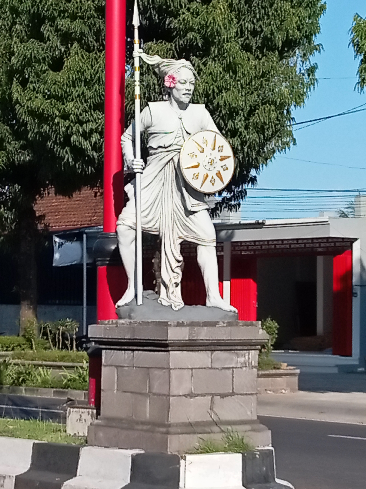 A statue of a warrior, possibly historical or legendary, stands in the foreground of the image. The warrior is depicted in a dynamic pose, holding a spear in one hand and a shield in the other. The statue is made of stone and is adorned with a pink flower on its head. The statue is placed on a stone pedestal. In the background, there are trees, buildings, and a blue sky. The scene appears to be an outdoor public space, possibly a monument or a landmark in the city of Blahbatuh, Indonesia, given the characteristic architecture of the building with red doors. The statue and its surroundings suggest a cultural or historical significance of the location.