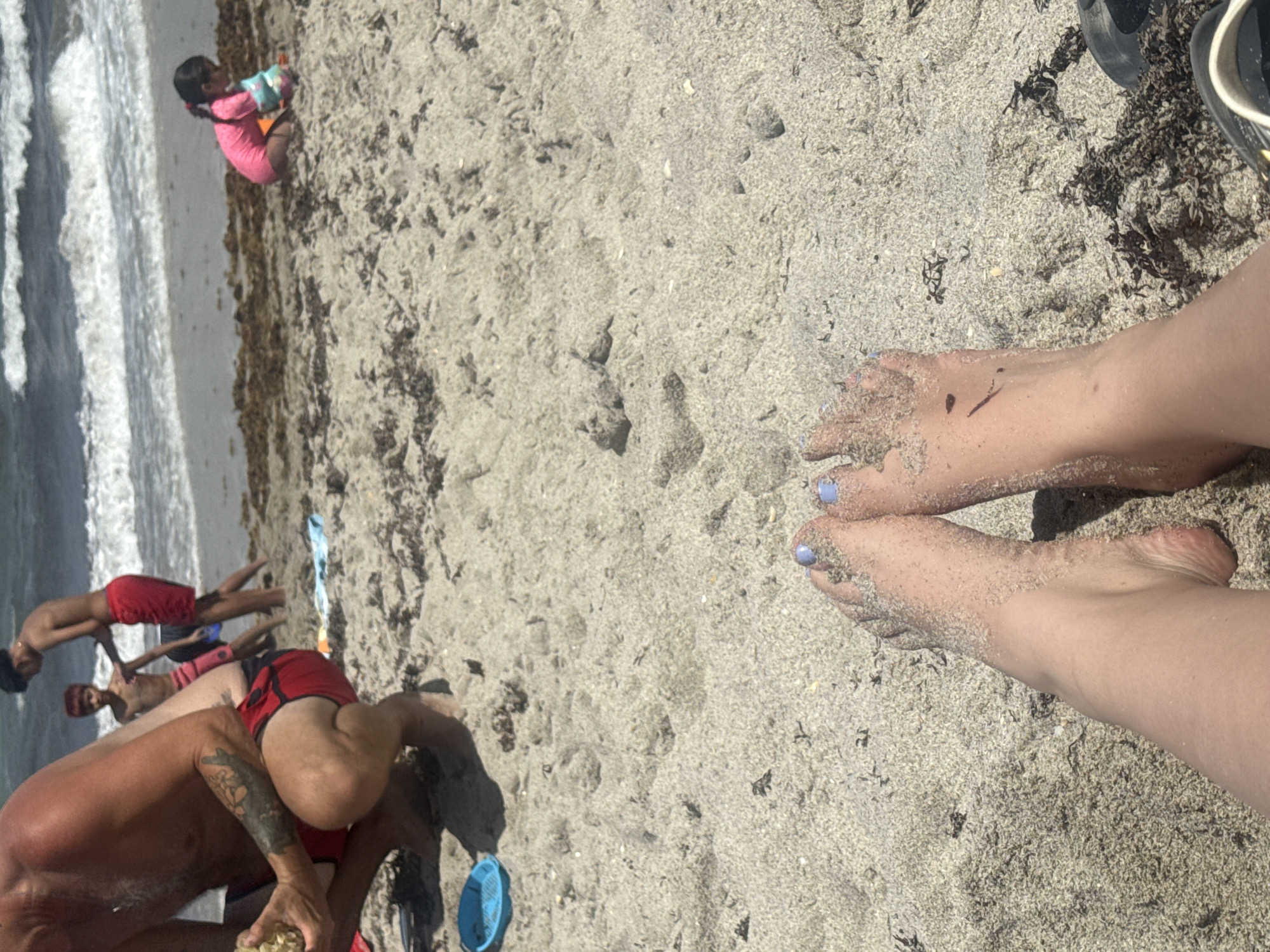 The image depicts a beach scene in Hollywood, Florida. In the foreground, a pair of feet with blue nail polish are covered in sand. Adjacent to the feet are flip flops, and seaweed. Further into the scene are several individuals of varying ages; one individual in a pink swimsuit near the ocean. More individuals who are mostly males with red shorts are also in the distance, closer to the foreground. In the background, waves are crashing along the shoreline. The sand is predominantly gray with some seaweed present.