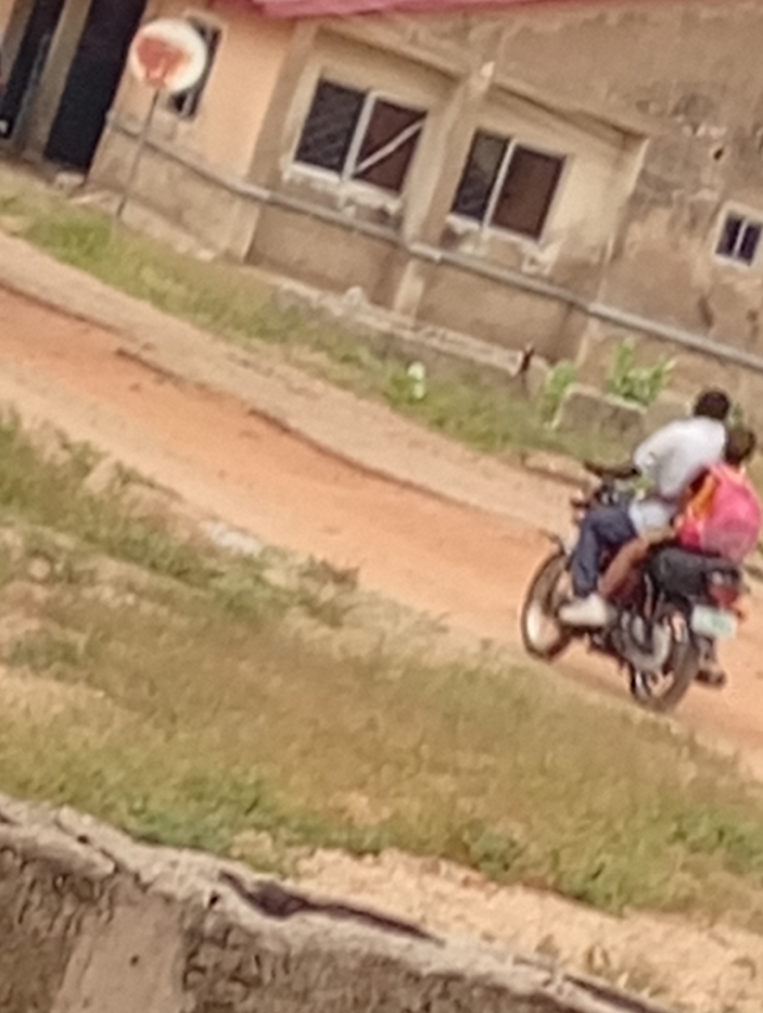 Two people are riding a motorcycle on a dirt road in front of a building. The passenger is wearing a pink backpack. The location is the city of Unknown, Unknown. The environment includes grass, a building with windows, and a pole with a circular object on top.