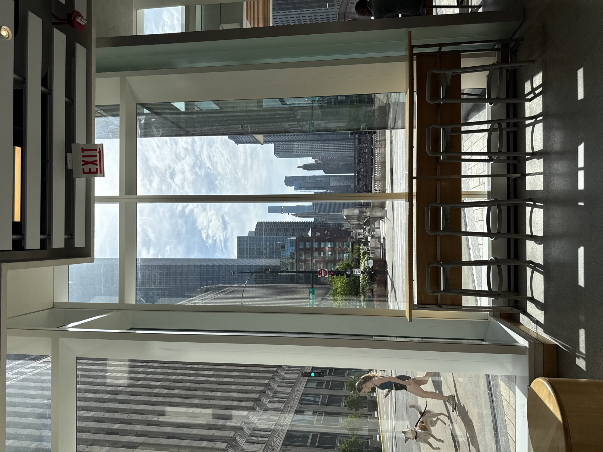 The image depicts a view from inside a building, looking out through a large window at a city street scene in Chicago, Illinois. In the foreground, there is a wooden counter with several bar stools. Through the window, a woman is walking a dog on the sidewalk. The street scene includes buildings, traffic lights, street signs, and a bridge in the distance. An "EXIT" sign is visible on the ceiling inside the building. The overall atmosphere suggests an urban environment on a sunny day.