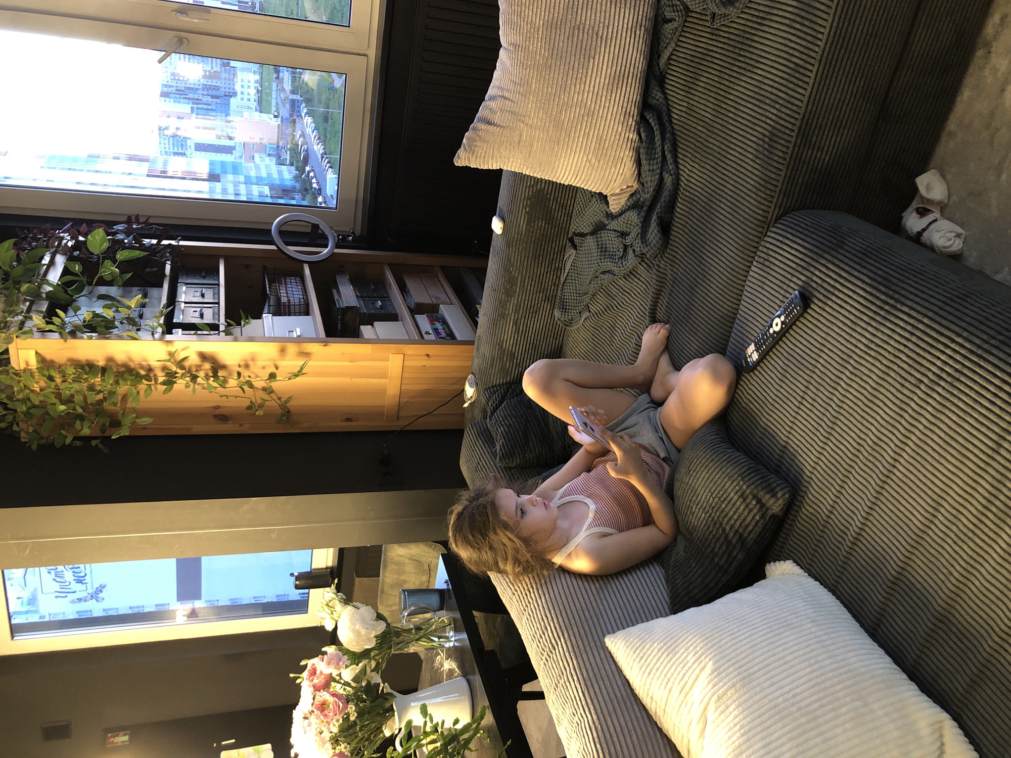 The image depicts a young girl sitting on a sectional sofa in a modern apartment, likely in Saint Petersburg, Russia, given the cityscape visible through the window. The girl is looking at her smartphone, holding it with both hands. She is wearing a pink and white striped tank top and gray shorts. The sofa is upholstered in a dark gray corduroy fabric and is adorned with several pillows, including a large white one on the left and a beige one on the right. A dark gray blanket is draped over the right side of the sofa.
To the left of the sofa, a dark dining table is partially visible with two vases of flowers and a blue glass. Behind the table, a window reveals a street or urban landscape with tall buildings. To the right of the girl, a tall, light-colored wooden bookshelf is filled with books and storage boxes. A ring light is attached to the side of the bookshelf, suggesting the area may be used for content creation. Further to the right, a large window offers a panoramic view of a modern cityscape with various buildings of different heights and colors, typical of Saint Petersburg. Cars are parked in a line outside. The overall lighting in the room is warm and ambient, with natural light entering from the windows. A television remote control rests on the armrest of the sofa. Underneath the sofa, a pair of rolled-up white socks with red accents are visible.